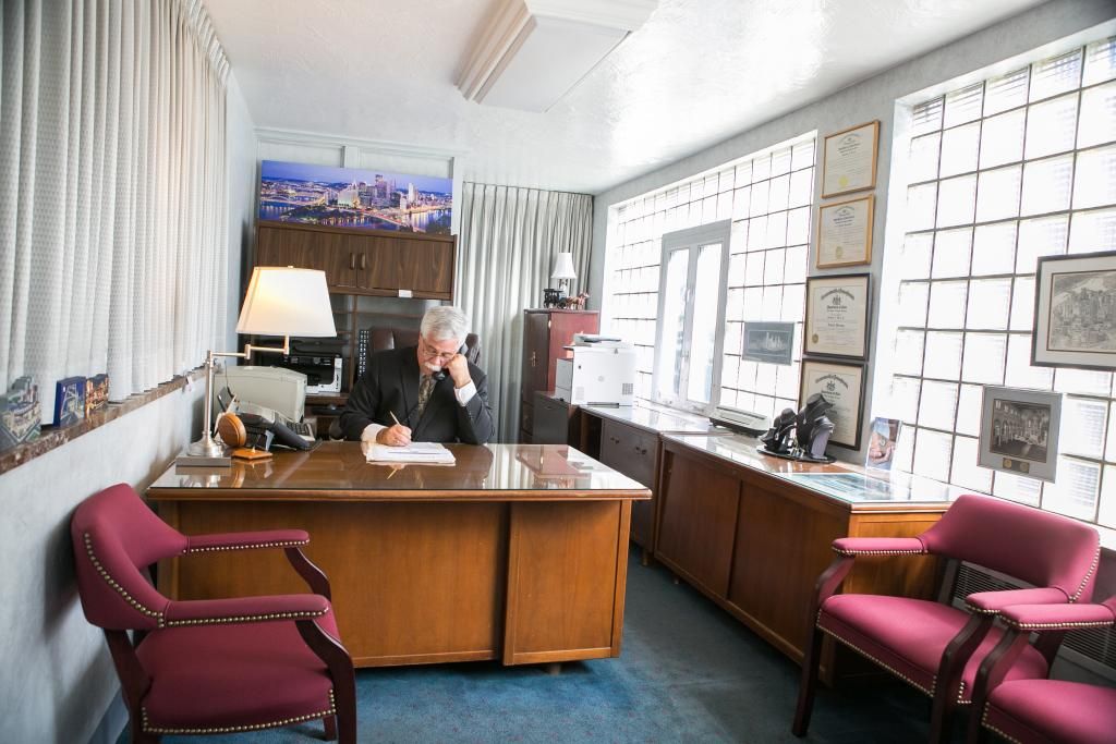 Man at desk in office, writing. Wooden desk, maroon chairs, glass block window.