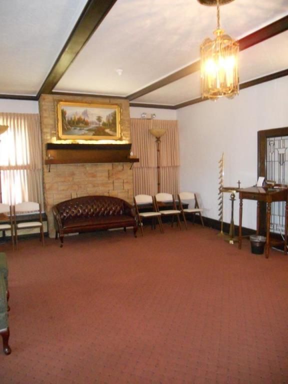 Funeral home interior: red carpet, sofa, chairs, fireplace with painting, and ornate lighting.