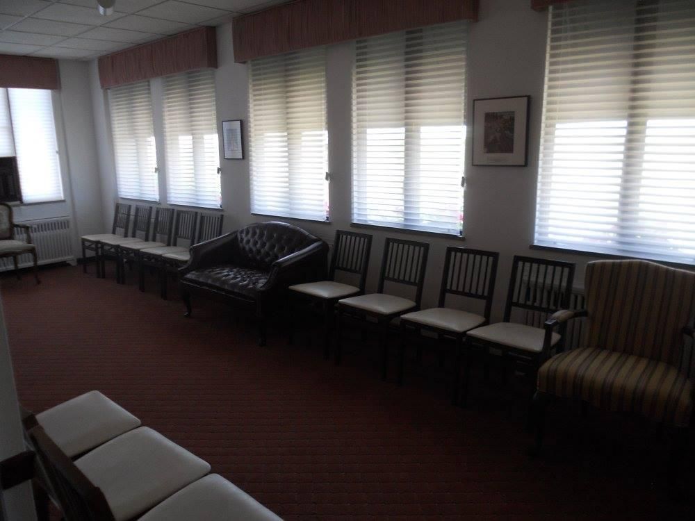 Waiting room with multiple chairs arranged near windows. Red carpet and white walls.