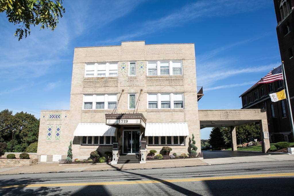 Three-story beige brick building with awning over entrance and connected walkway; sunny day.