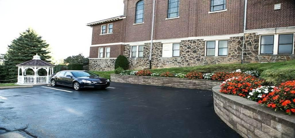 A dark car parked in front of a brick building with a gazebo and flowerbed. Cloudy sky.