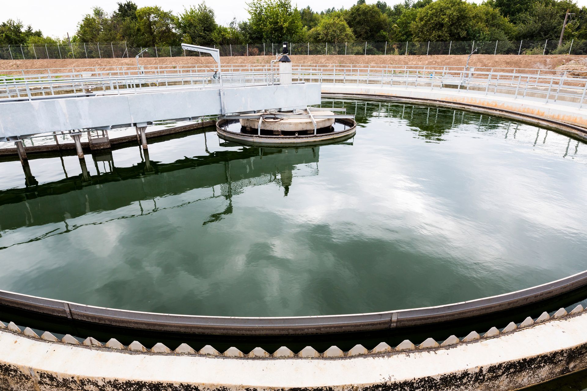 Large circular water treatment tank with a calm green surface and a central platform, outdoors under cloudy sky