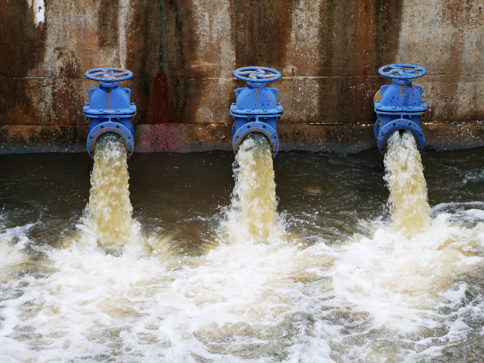 Three blue water valves releasing strong streams into a churning river or canal