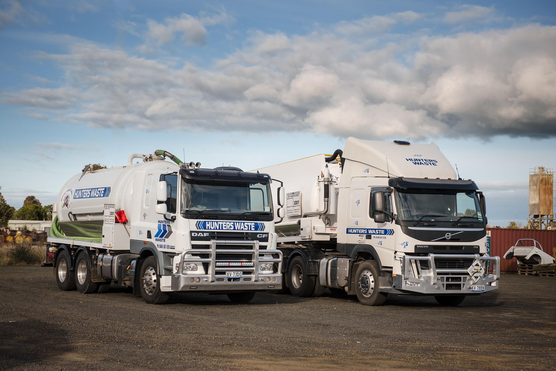 Two white garbage trucks parked side by side on a gravel lot under a cloudy sky