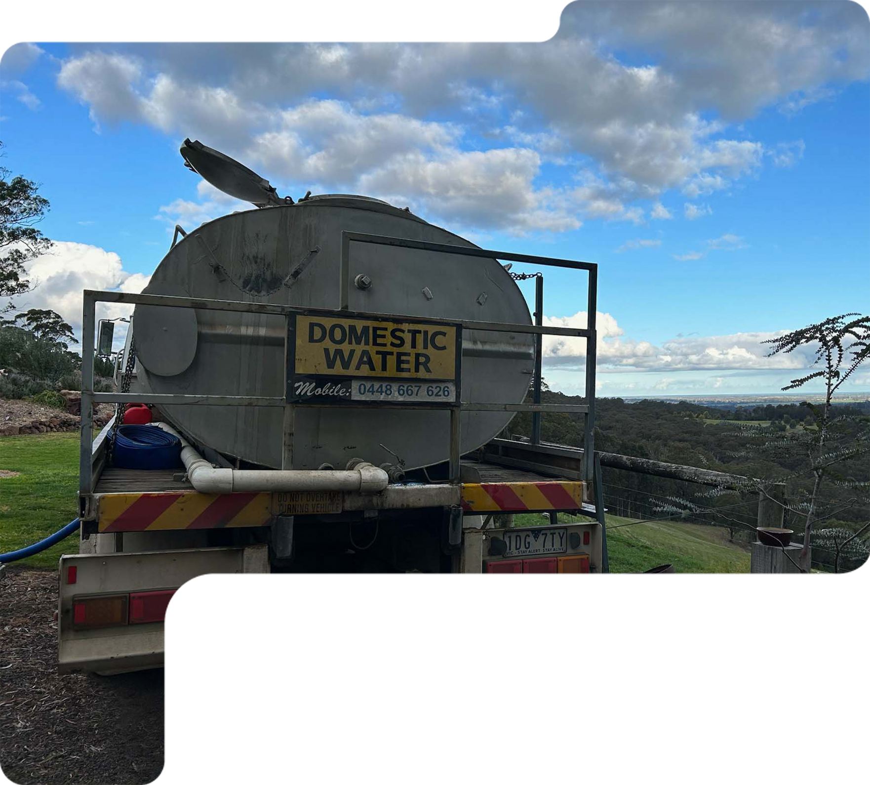 Water tanker truck with “Domestic Water” sign on a gravel road by the coast under a cloudy blue sky
