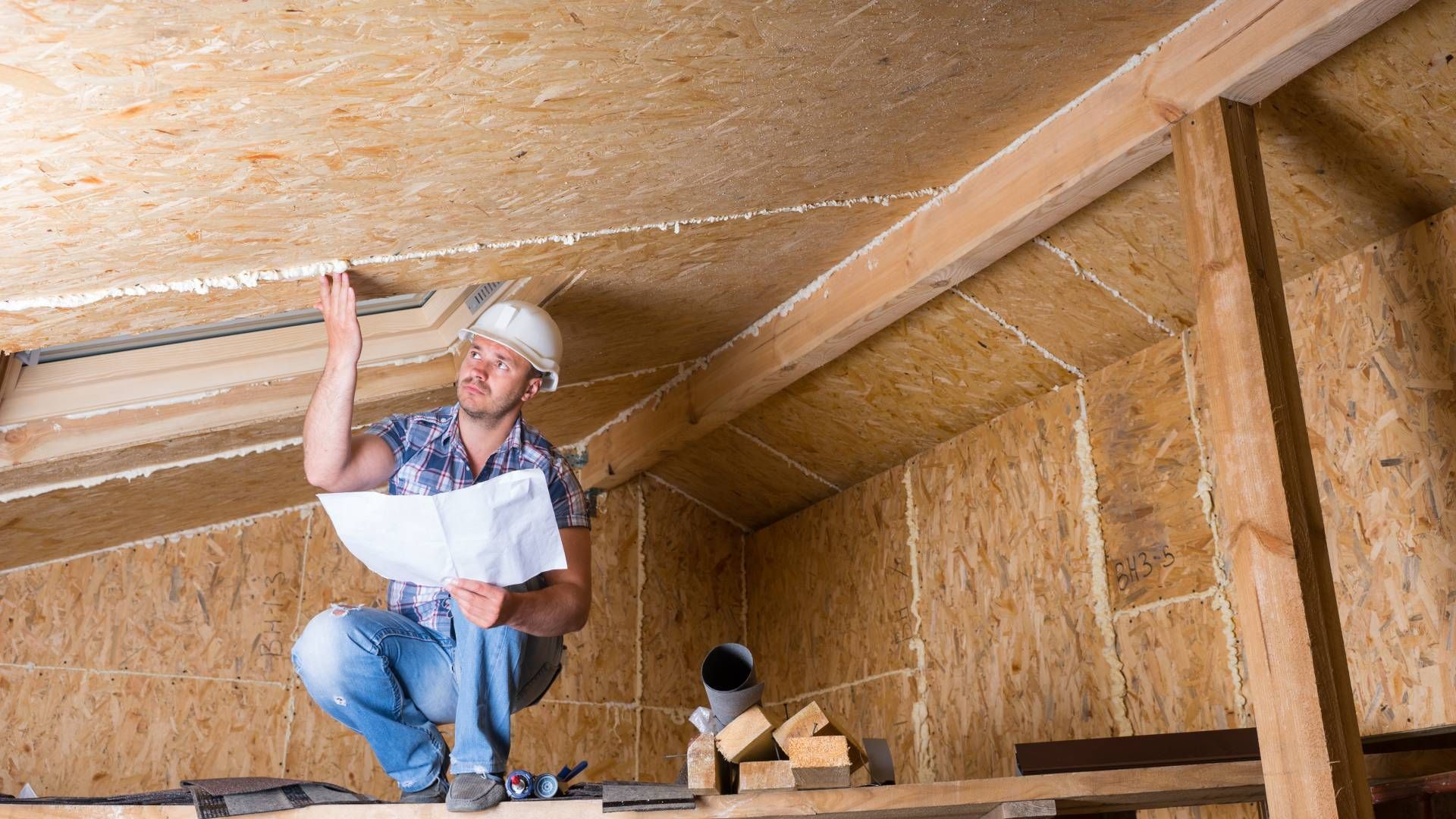 Construction worker inspecting attic insulation. Holding blueprints, wearing a hard hat, pointing upwards.