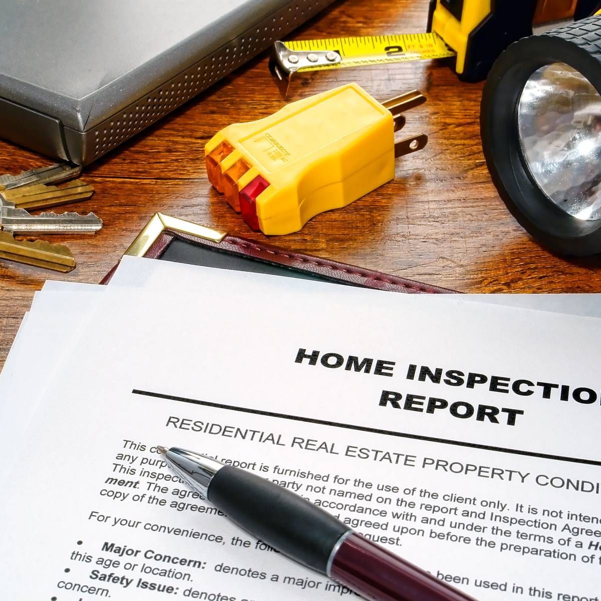 Home inspection report with pen, keys, measuring tape, flashlight, and adapter on a wooden surface.