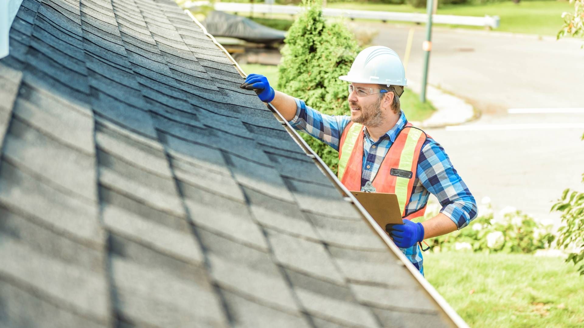 Roofer inspecting a dark gray shingled roof, wearing a hard hat, safety vest, and gloves. Holding a clipboard, outdoors.