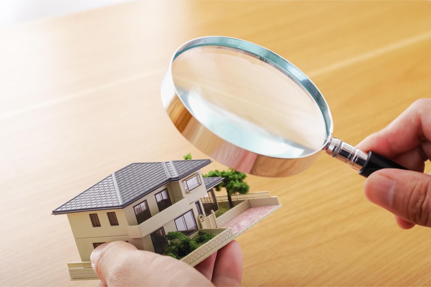 Hand holding a magnifying glass over a miniature house model on a wooden table.