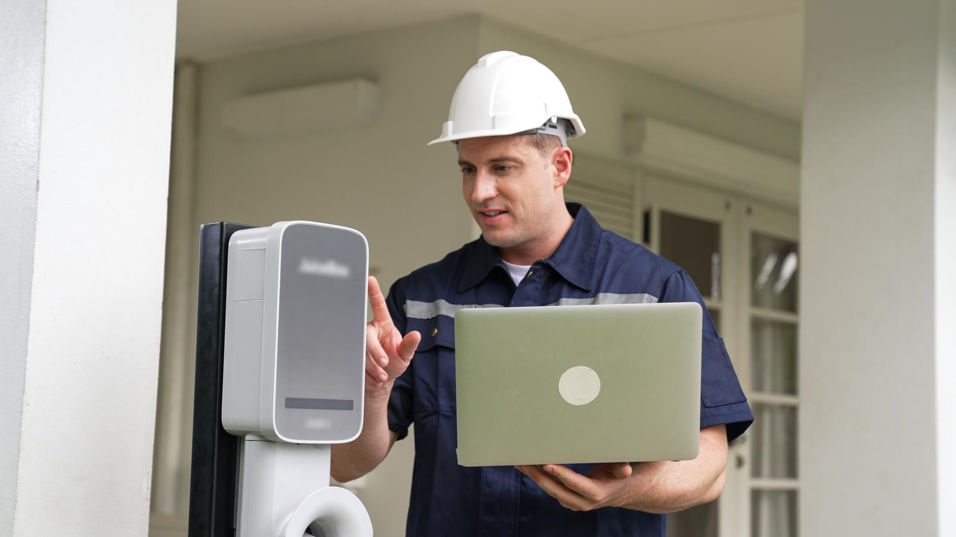 Man in a hard hat and work uniform, using a laptop, interacts with a device mounted on a pillar outdoors.