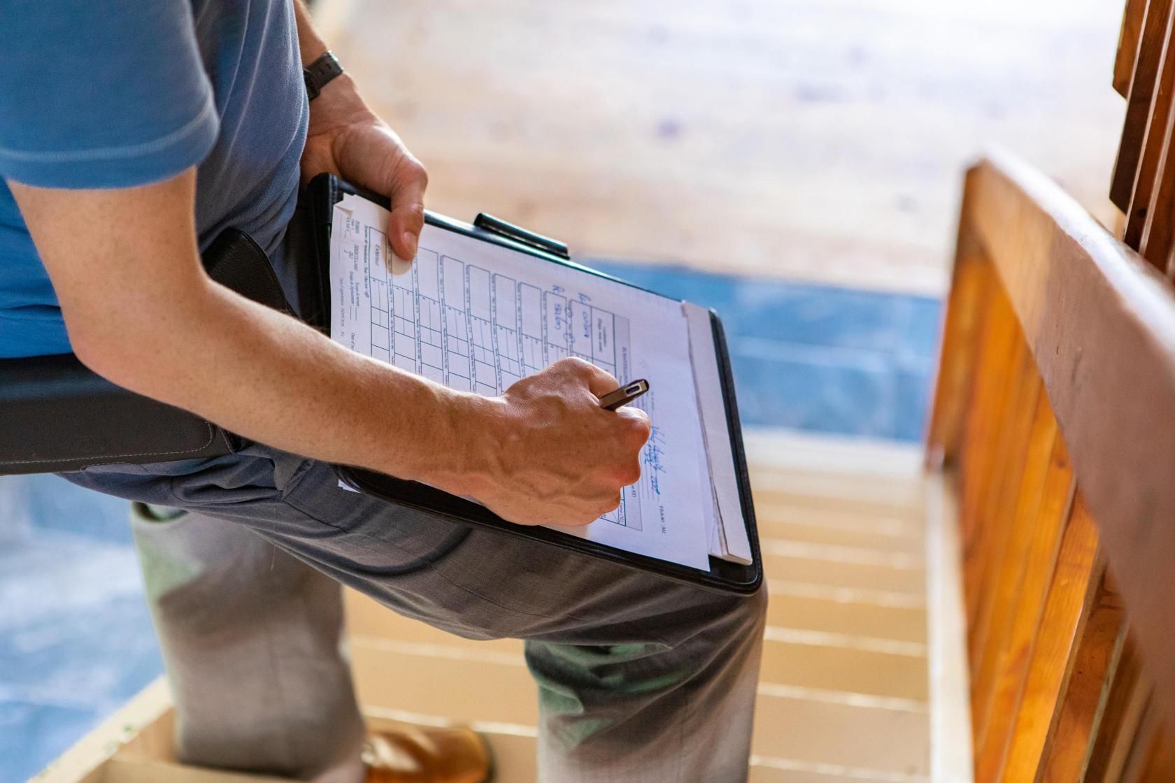 Person writing on a clipboard while standing on wooden stairs, inspecting the steps.