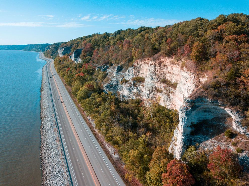 An aerial view of a highway next to a body of water surrounded by trees.