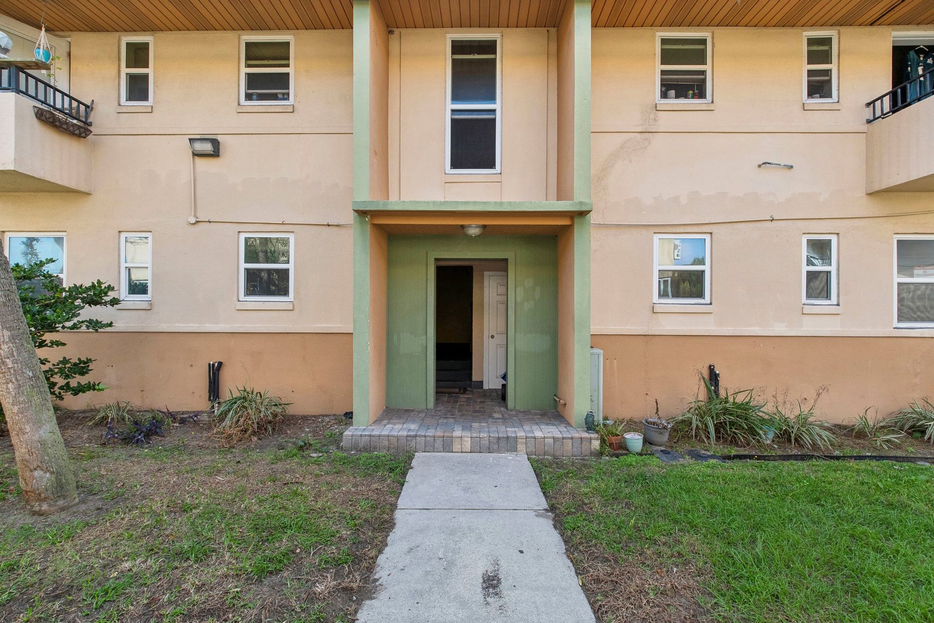 Two-story building exterior. Tan and green facade with windows and entrance. Concrete walkway and grass in front.