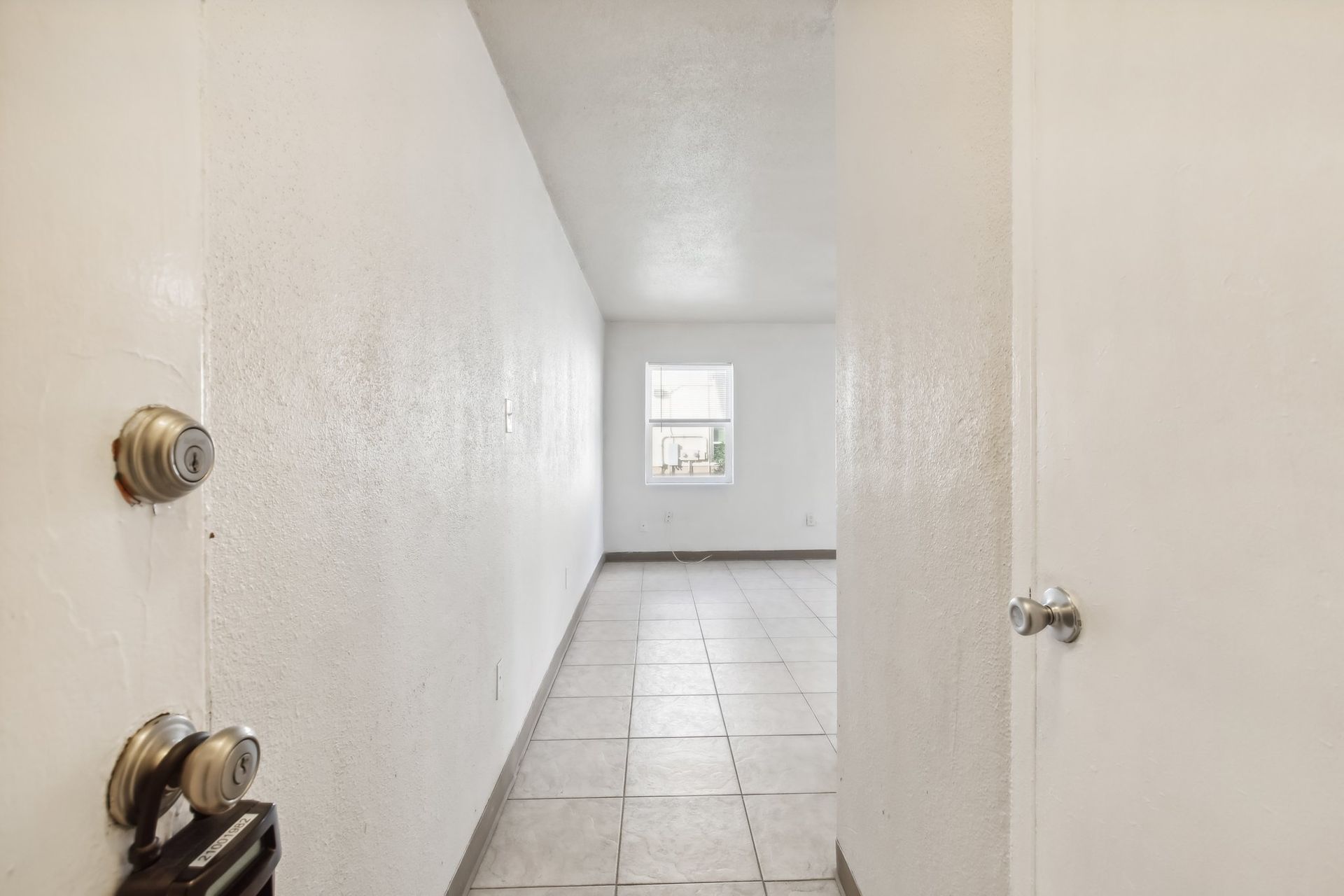 Narrow hallway with white walls and tiled floor, leading to a room with a window.