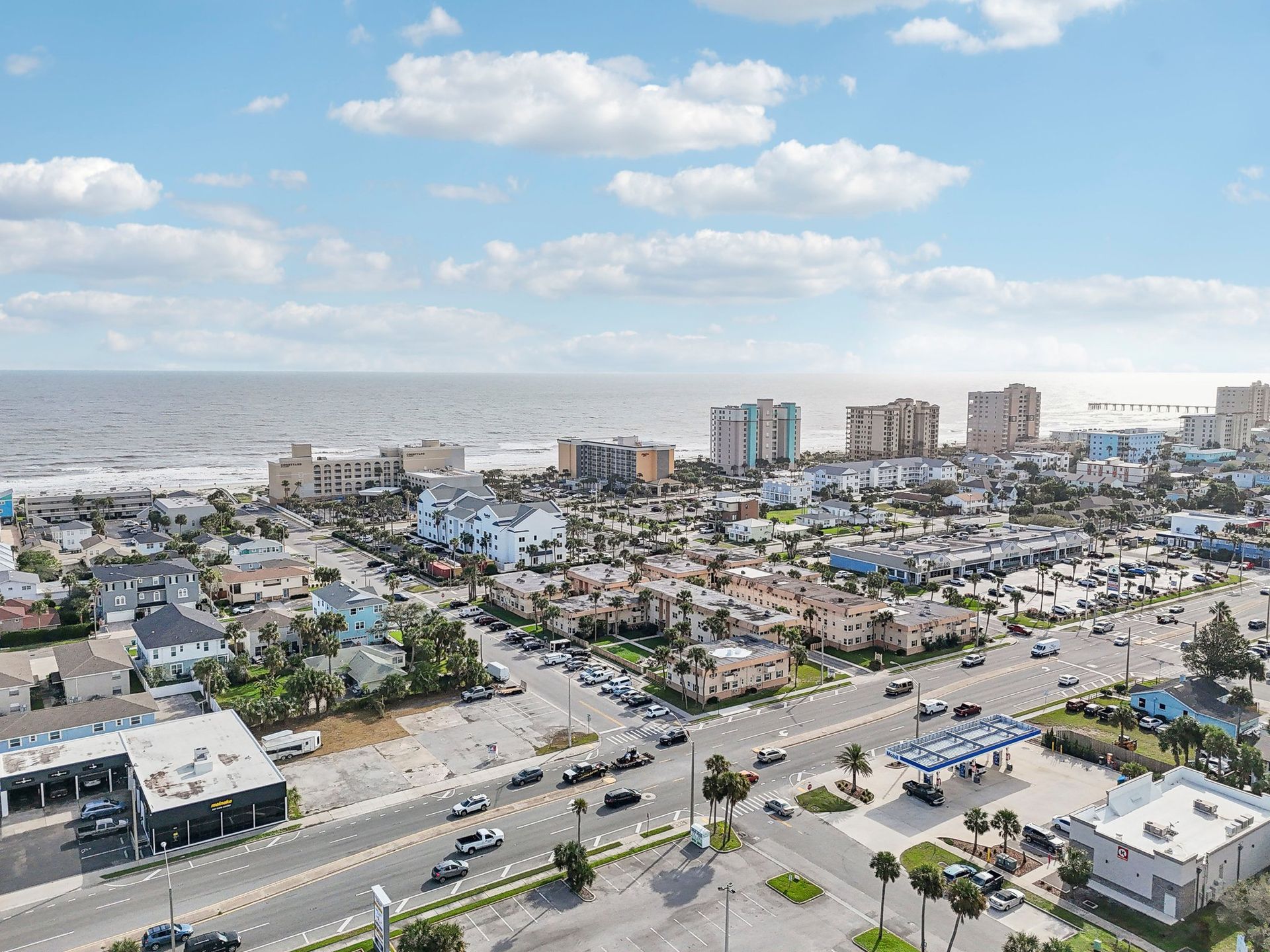 Aerial view of a coastal city with buildings, a road, and the ocean under a blue sky with clouds.
