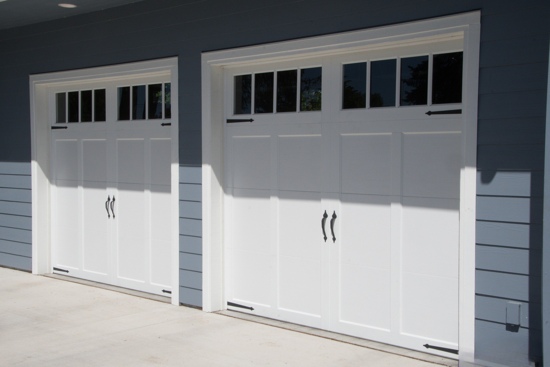 Two white steel carriage house garage doors with square windows and black handles on a gray home