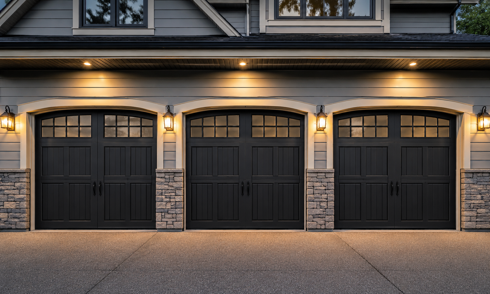 Three black steel carriage house garage doors with arched window panes, stone accents, and illuminated sconce lighting above