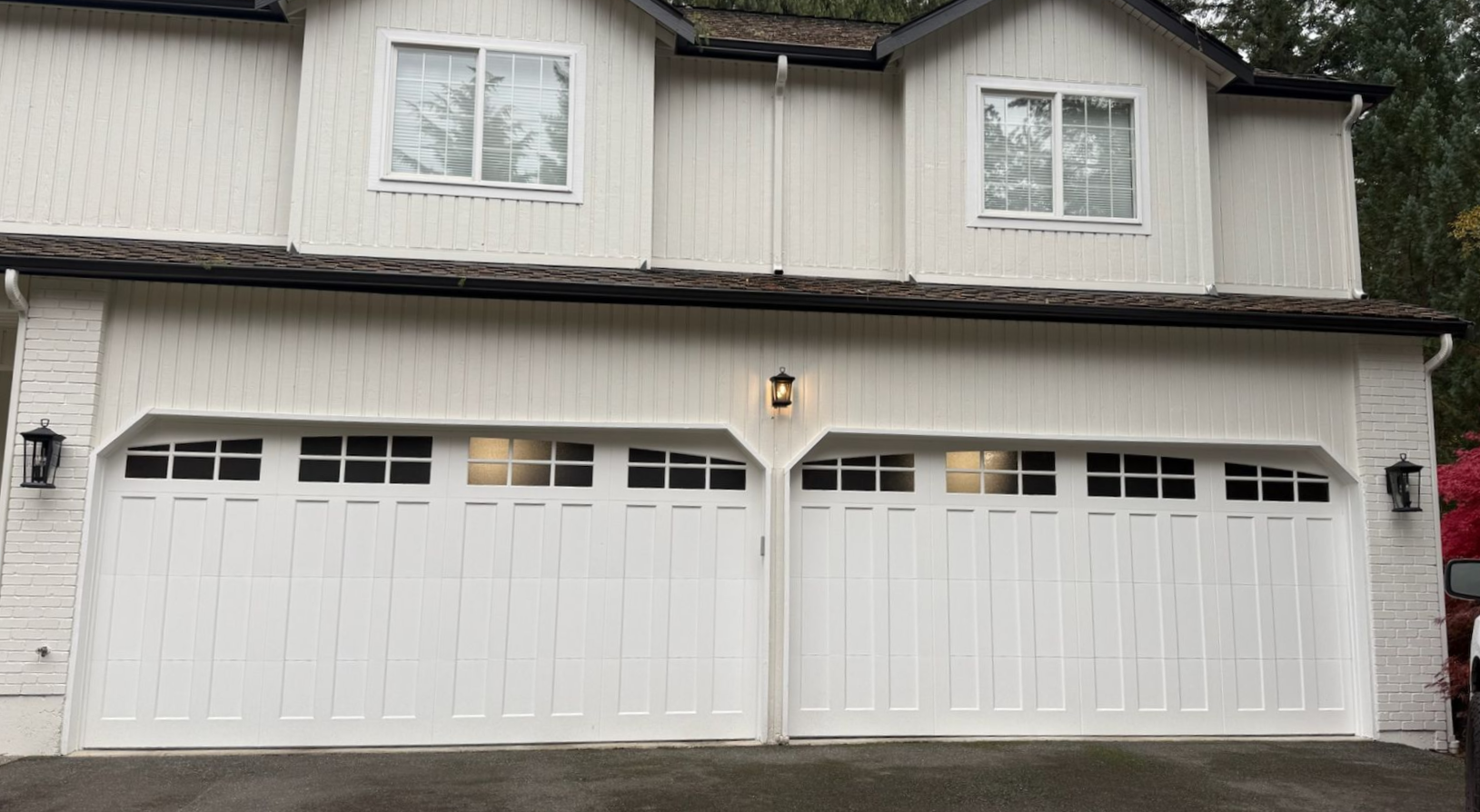 White home with two white steel carriage house garage doors, square window panes, and wall sconce lighting between the doors
