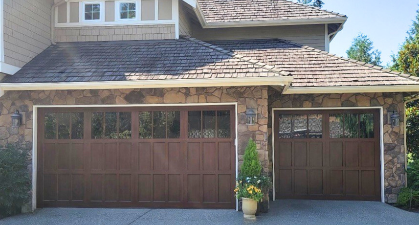 Two dark wood-tone steel carriage house garage doors with square windows on a stone-accented home