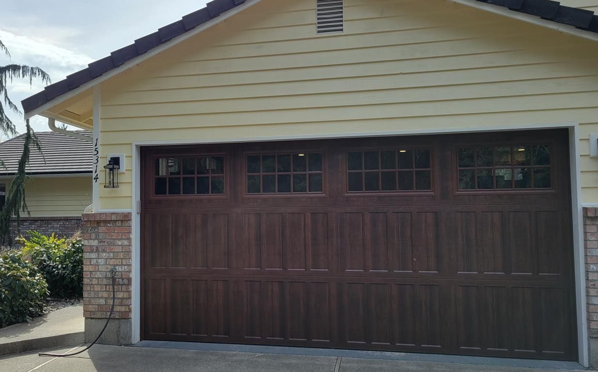 Tan home with a wide dark wood-tone steel carriage house garage door, square window panes, and a stone column base