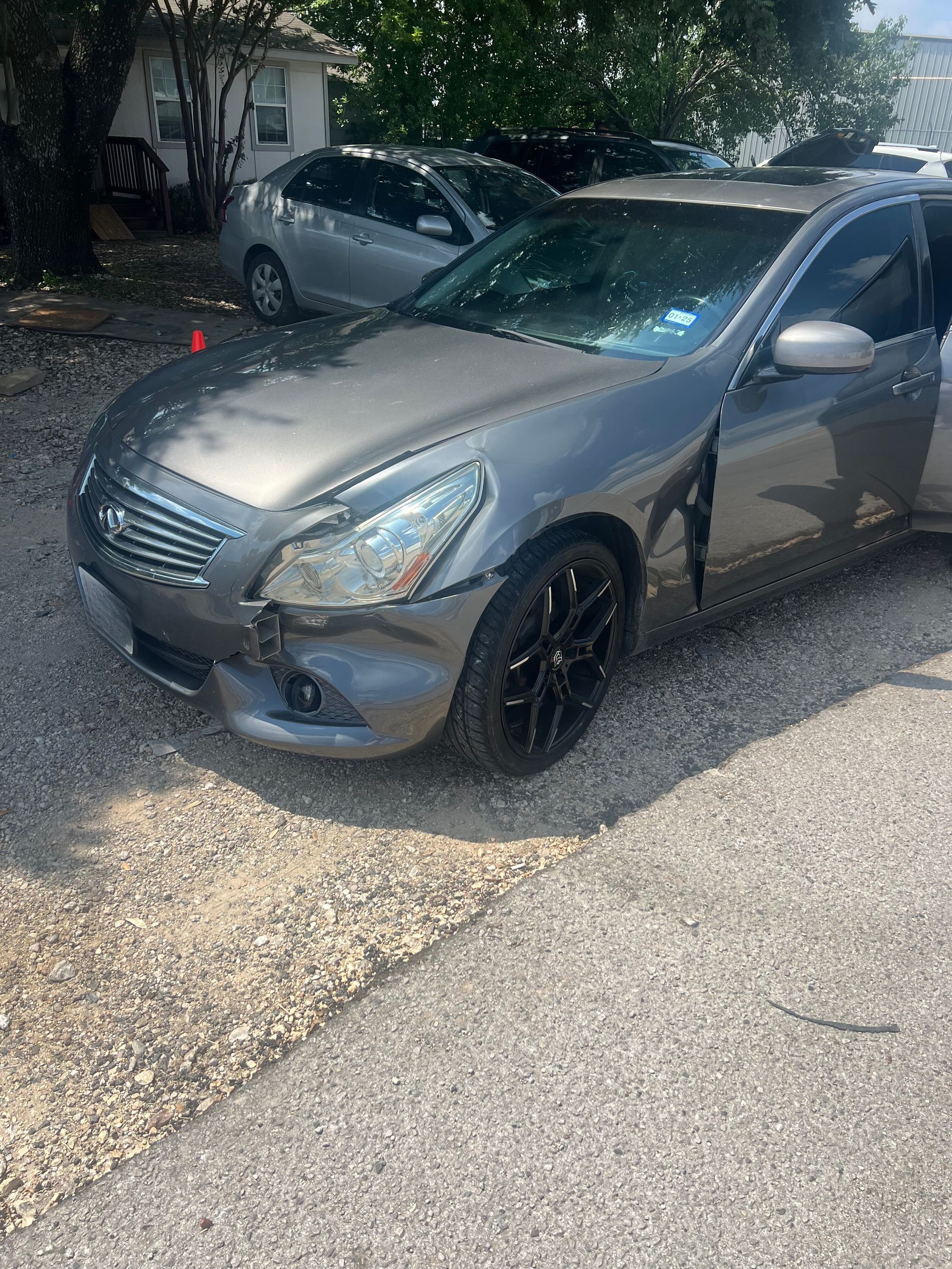 A gray car with a damaged bumper is parked in a gravel lot.