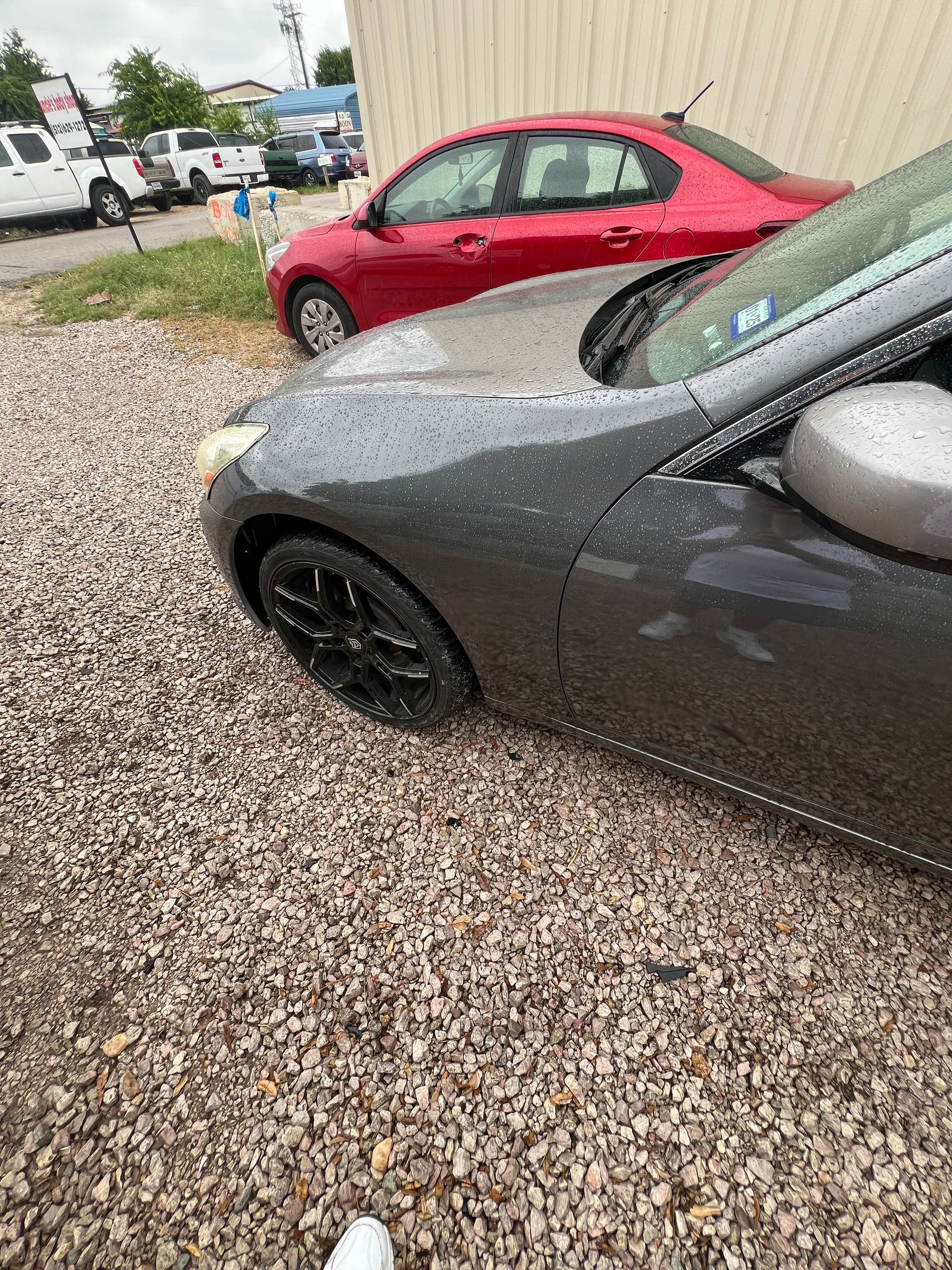 A gray car is parked next to a red car in a gravel lot.