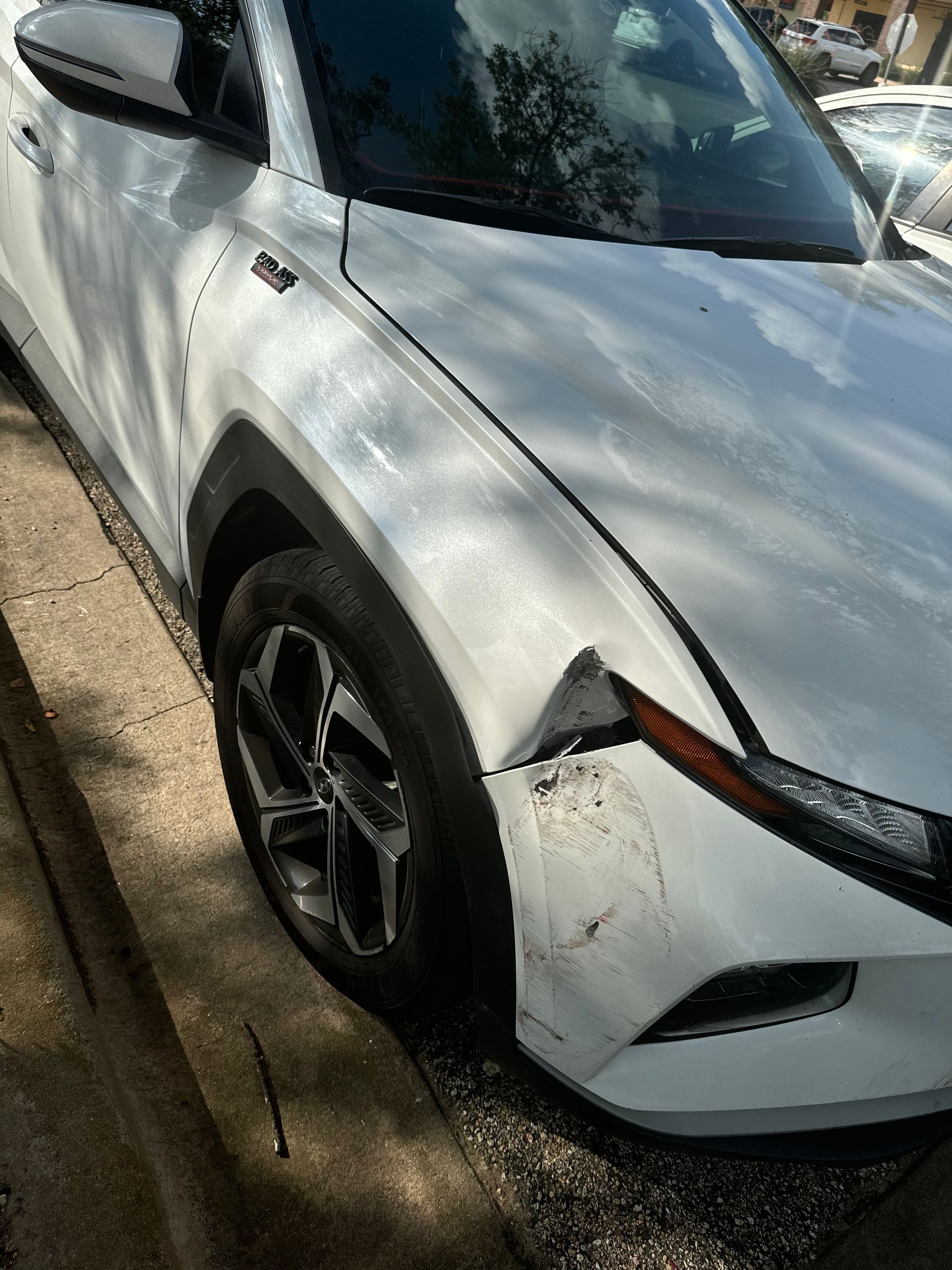 A white car with a damaged fender is parked on the side of the road.