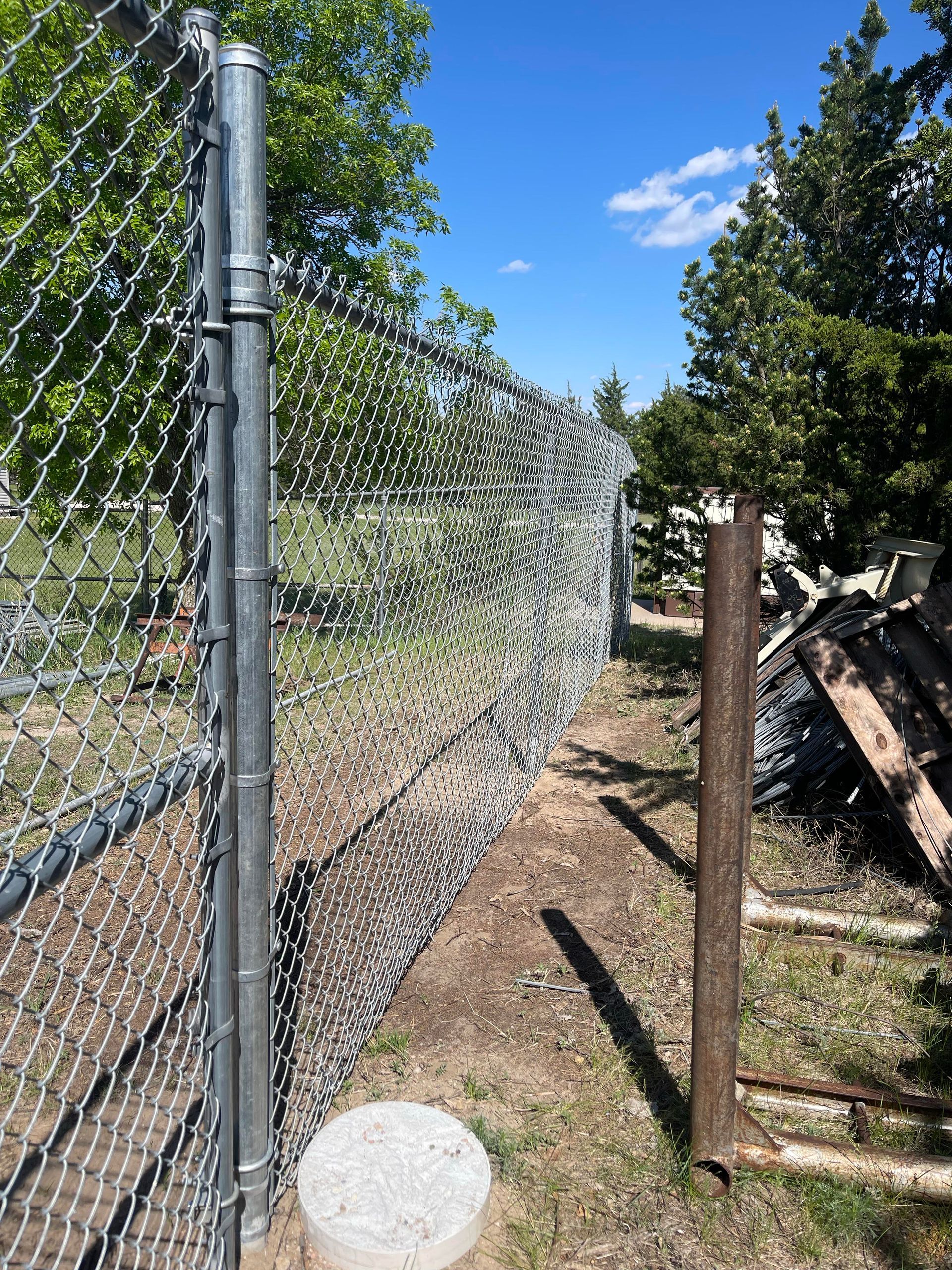 Chain-link fence along a gravel path beside trees and a pile of wood under a blue sky.