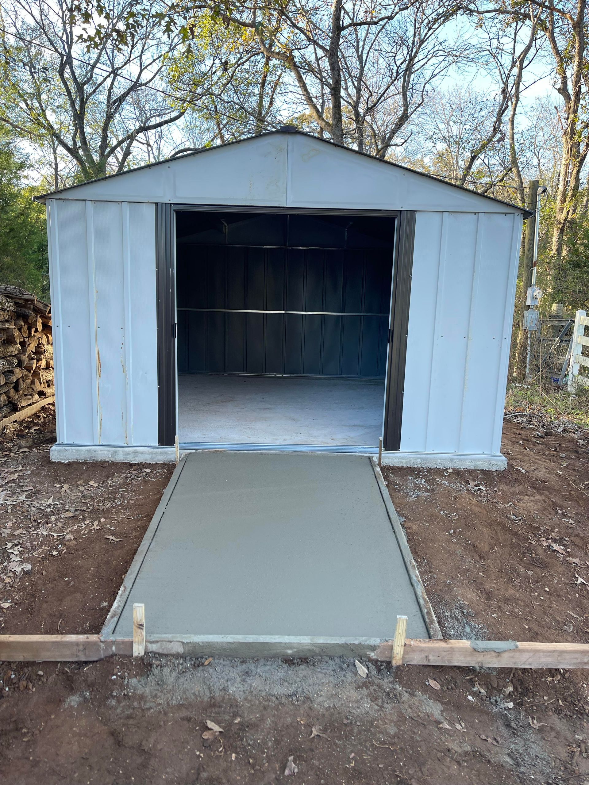 White metal shed with open doorway and a concrete ramp leading to it, outdoors among trees