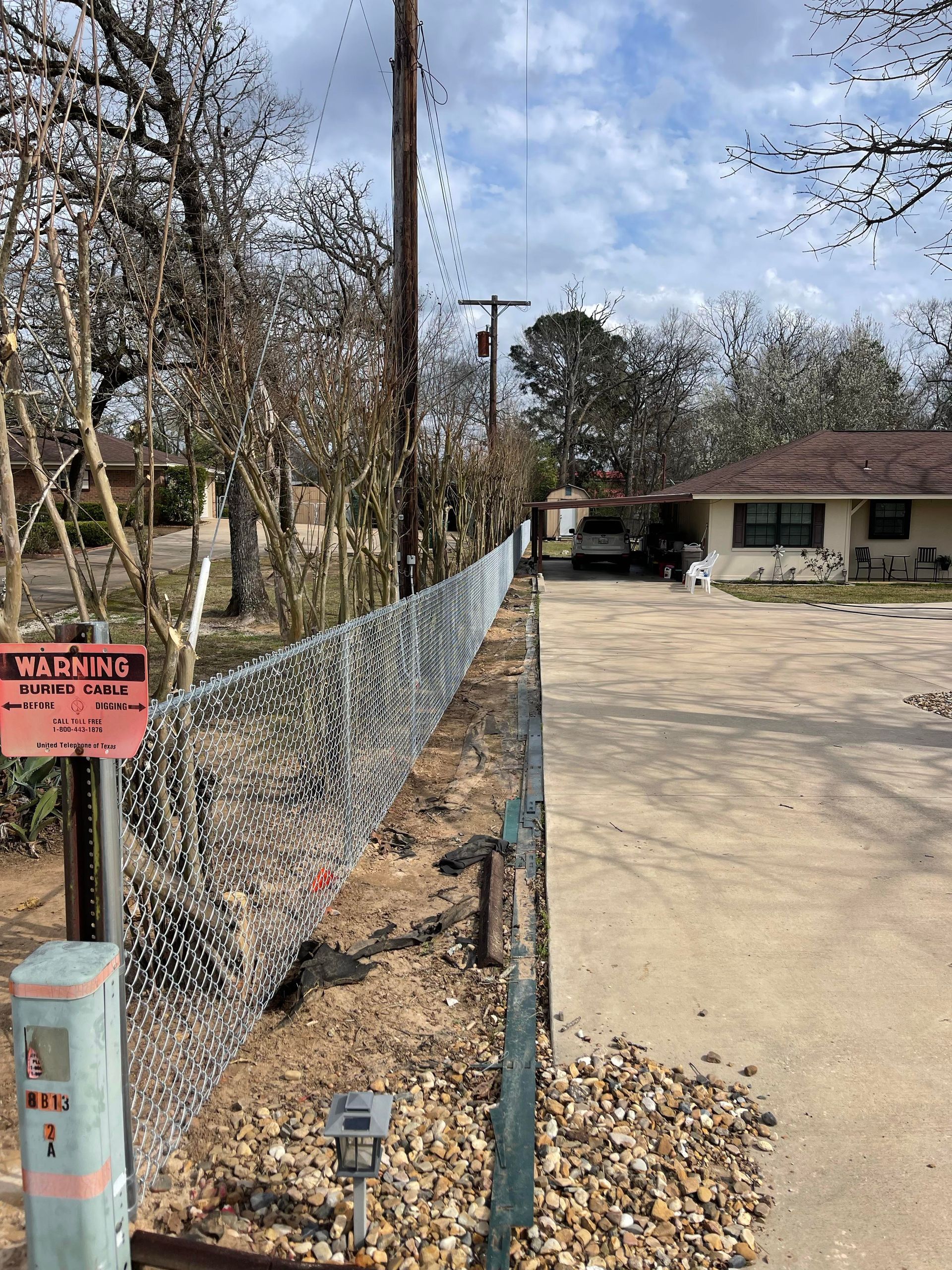 Chain-link fence beside a dusty driveway, with a warning sign and trees lining the property.