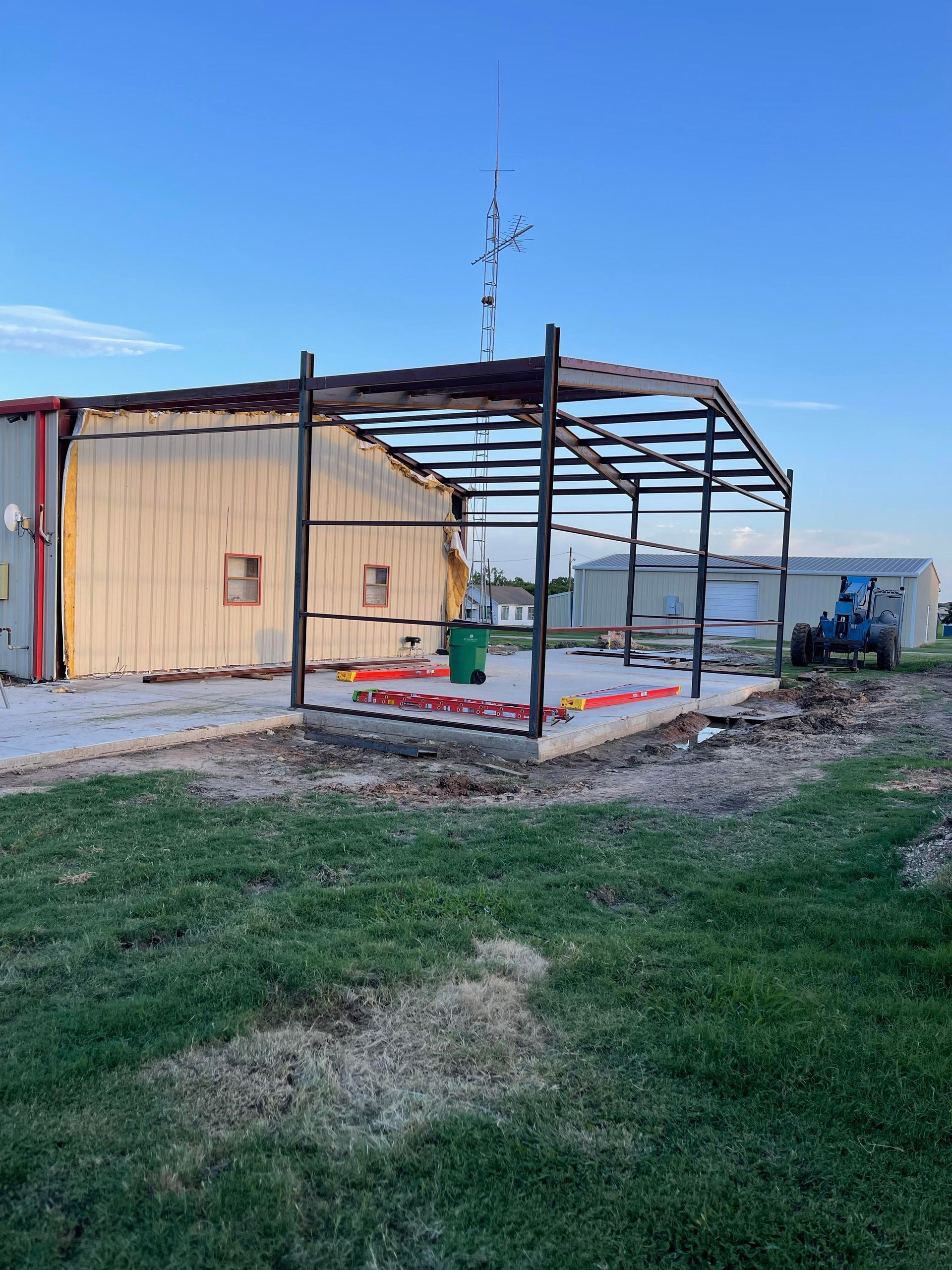 Partially built metal shed beside a tan building on a grassy lot under a clear blue sky