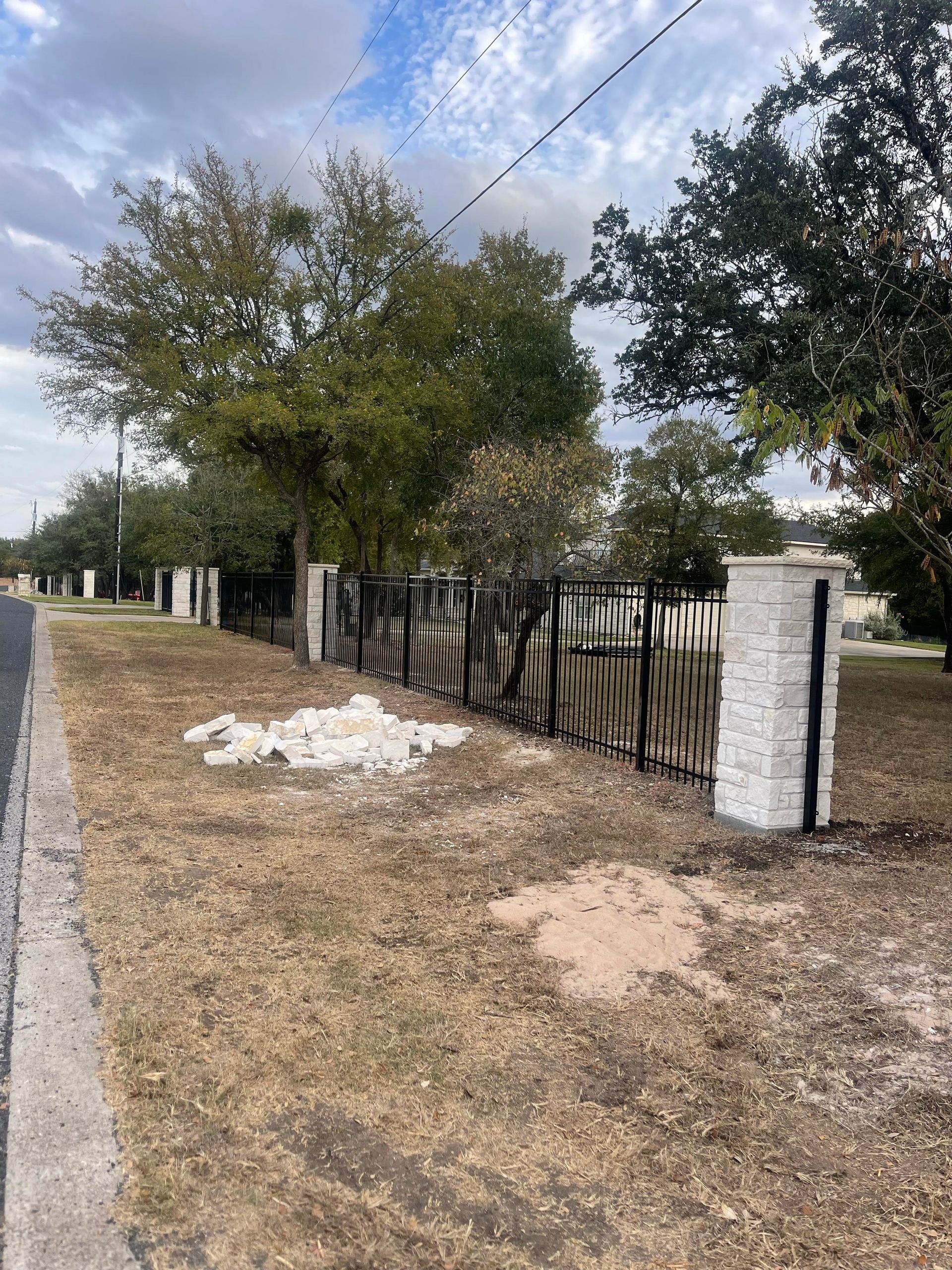 Quiet roadside with a wire fence, white posts, dry grass, and trees under a cloudy sky.