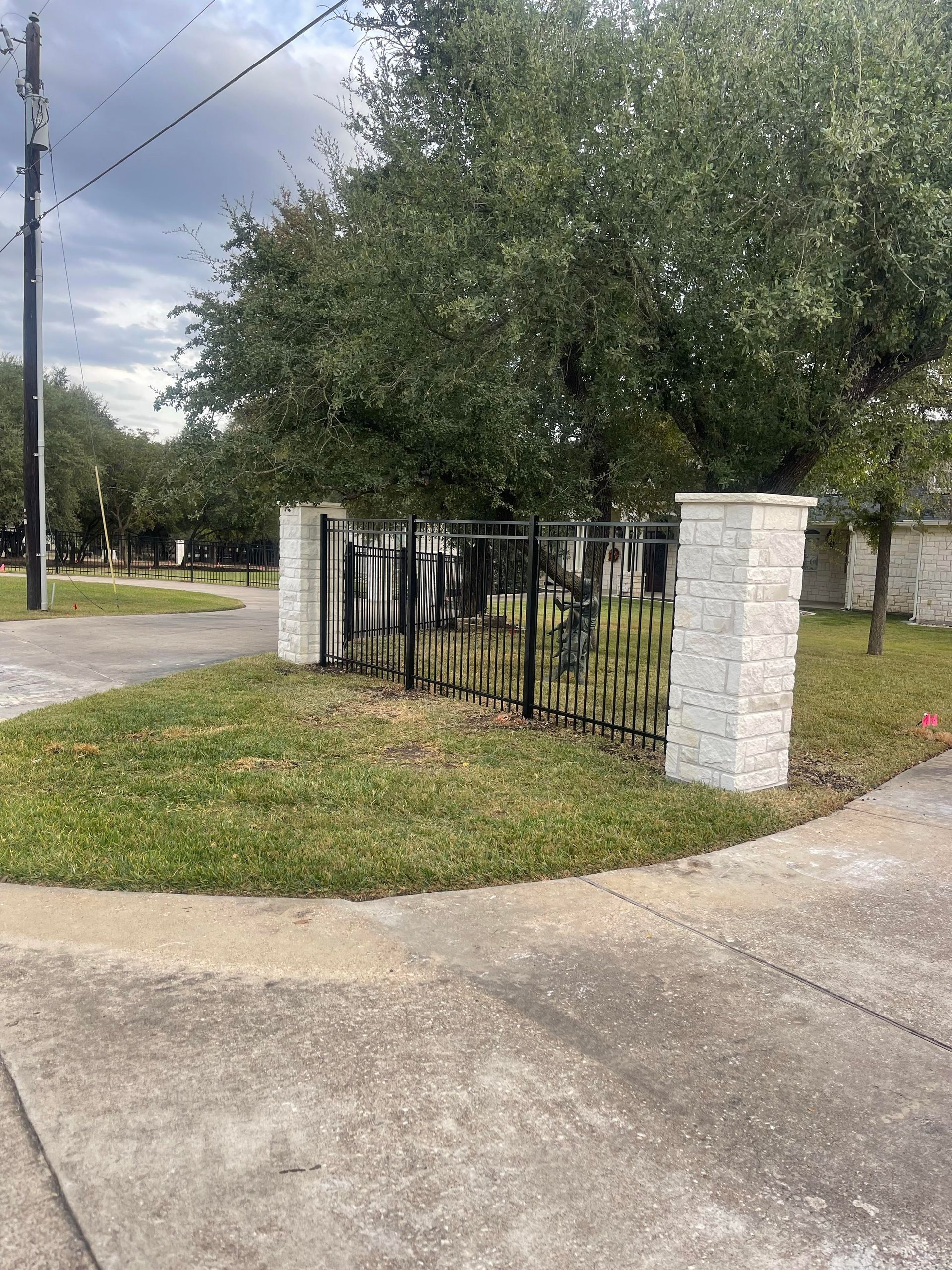 Corner lot with black iron fence and stone pillars beneath a large tree beside a road.