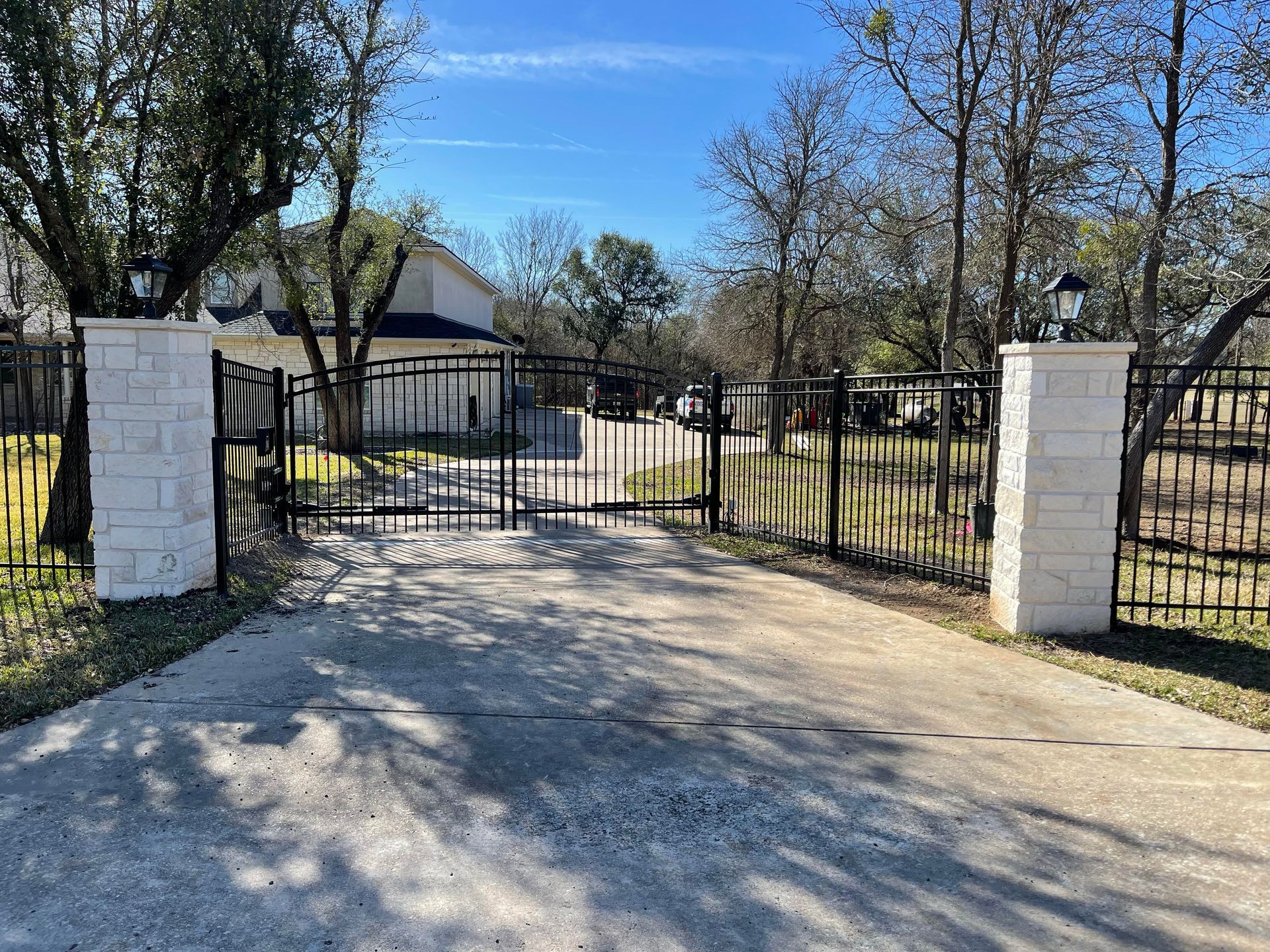 Black metal gate between white brick pillars opening to a driveway and house in a sunny yard