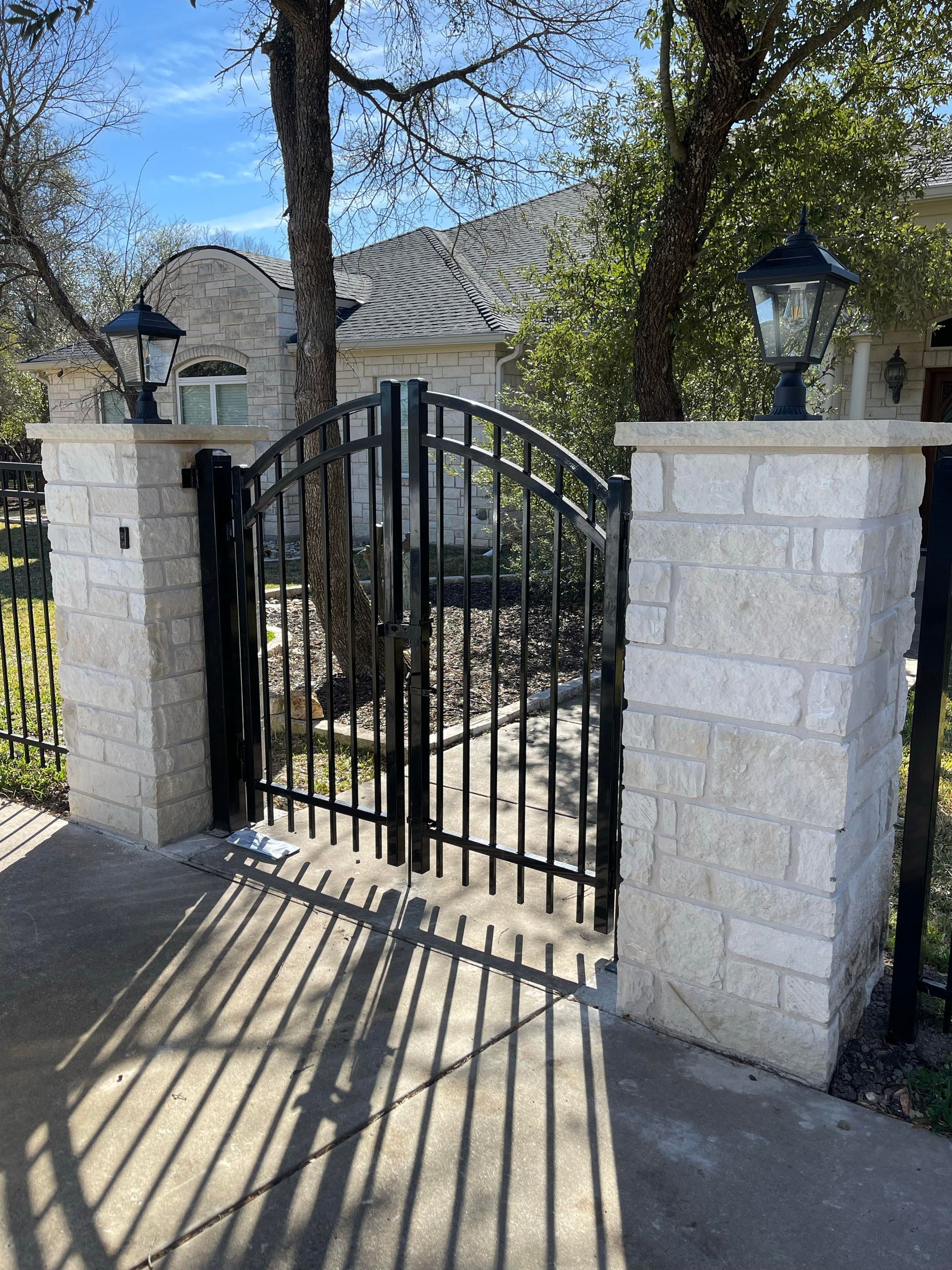 Black metal gate between stone pillars at a house entrance, with tree shadows on the driveway.