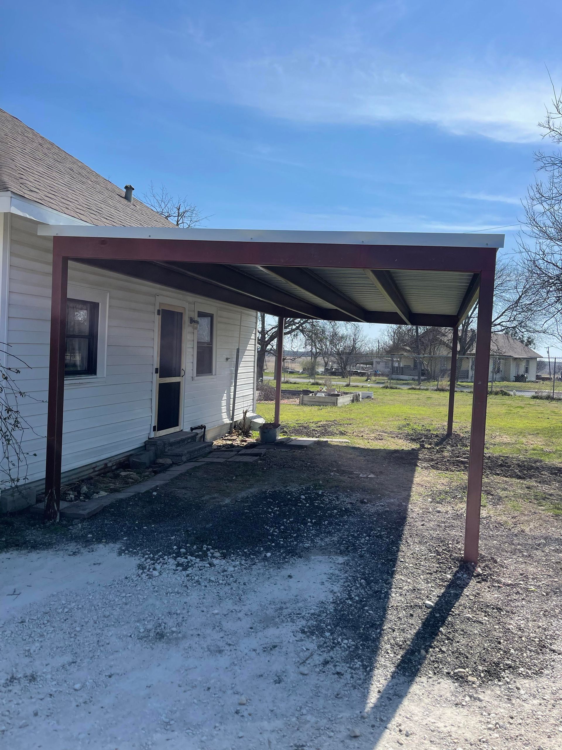 White house exterior with a rusted metal carport and gravel driveway under a blue sky