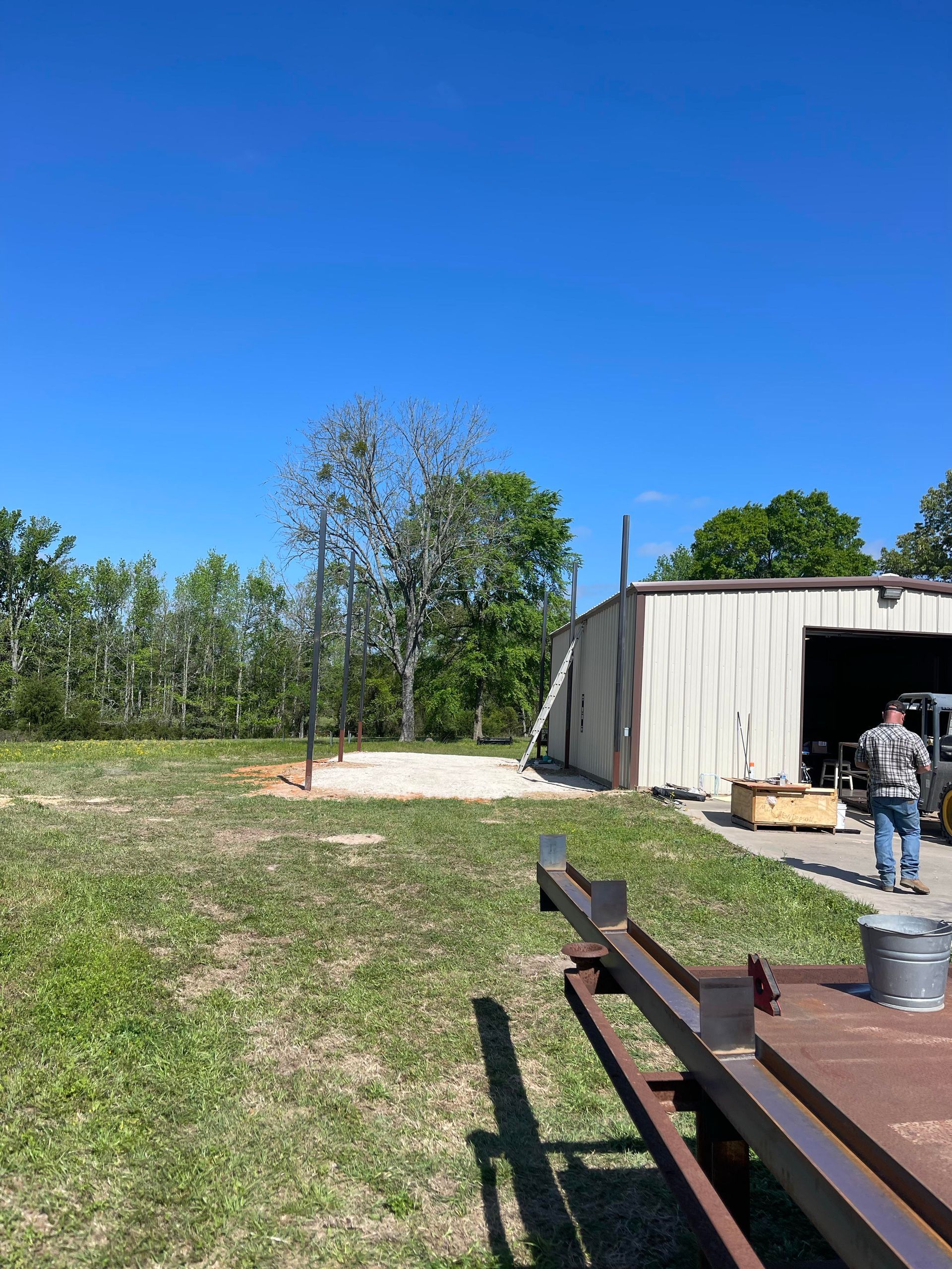 Outdoor yard with a metal shed, grassy field, trees, and a person near an open doorway beside a picnic table.