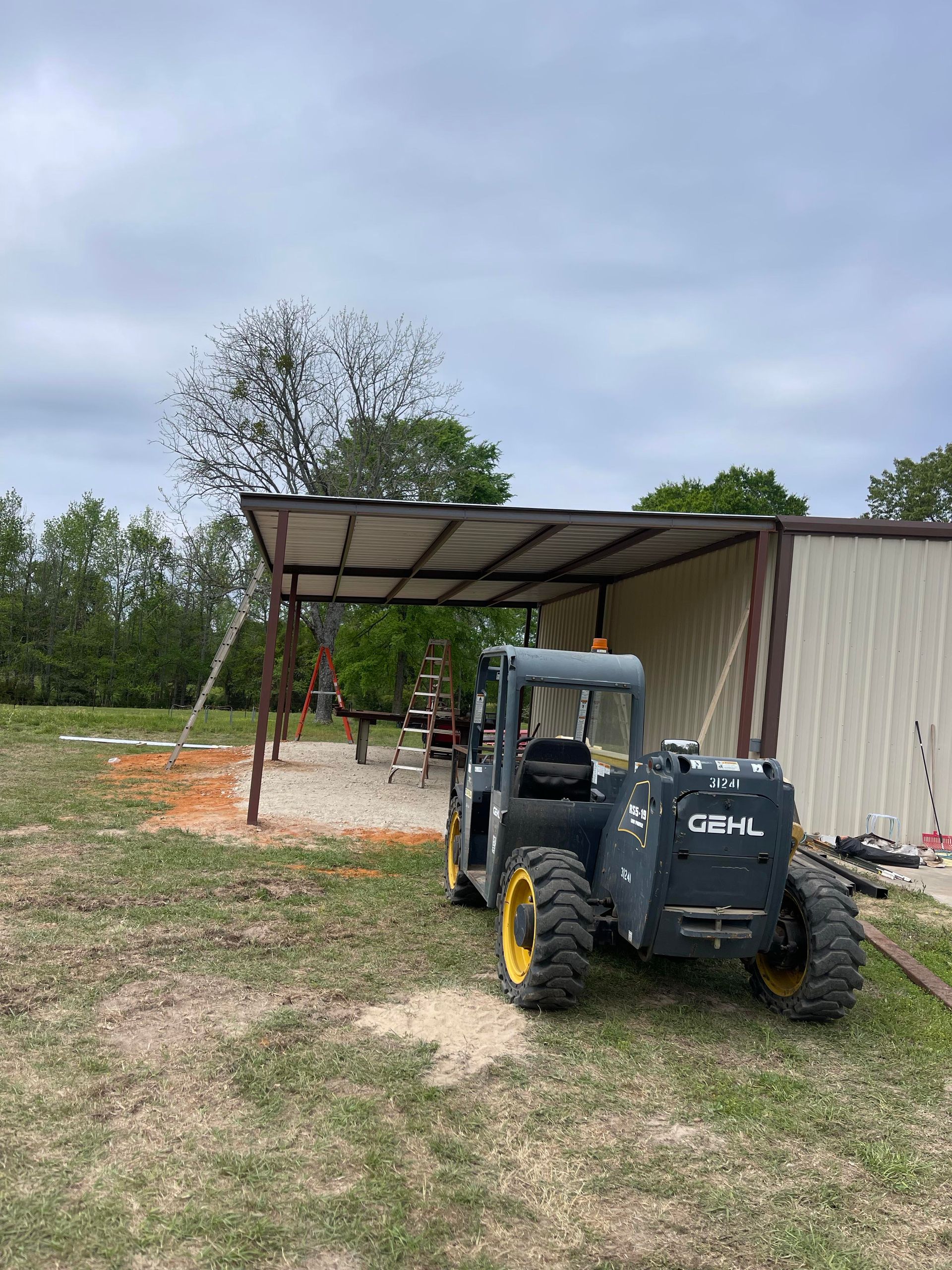 Black tractor parked beside a metal shed and carport on a grassy rural property.