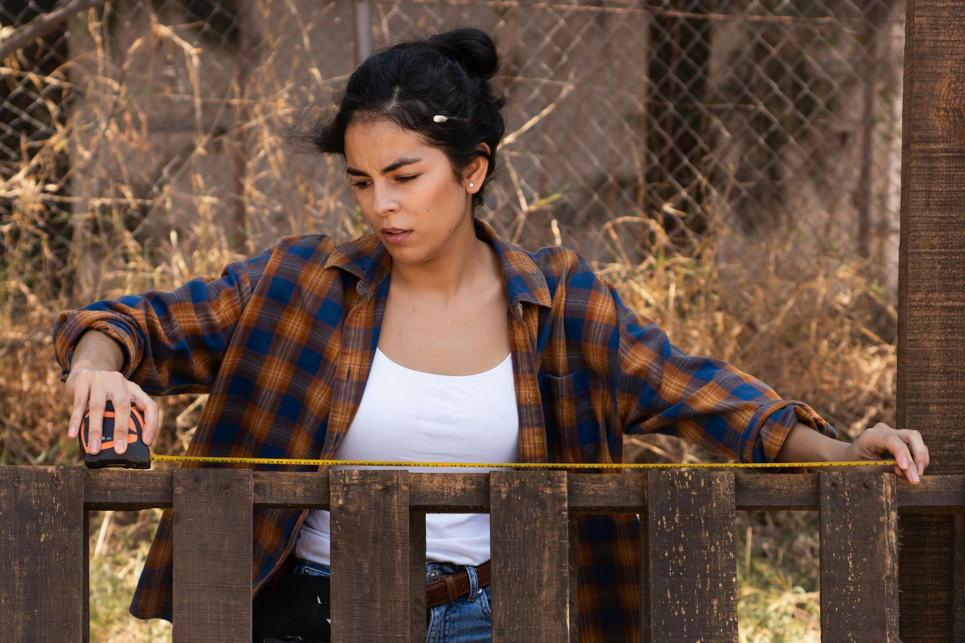 Woman in a plaid shirt leaning on a wooden fence outdoors, looking down thoughtfully.