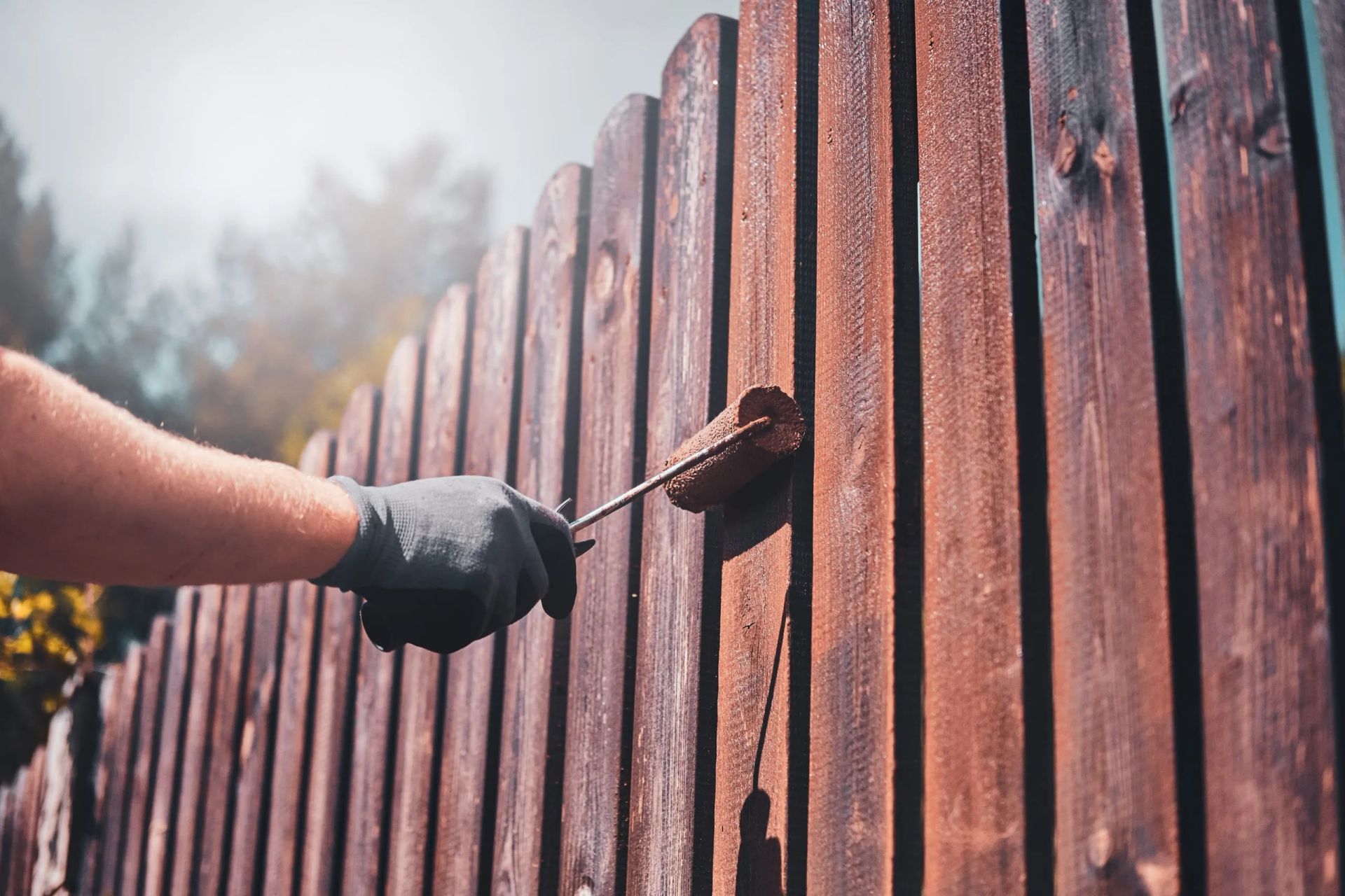 Gloved hand painting a brown wooden fence with a brush outdoors