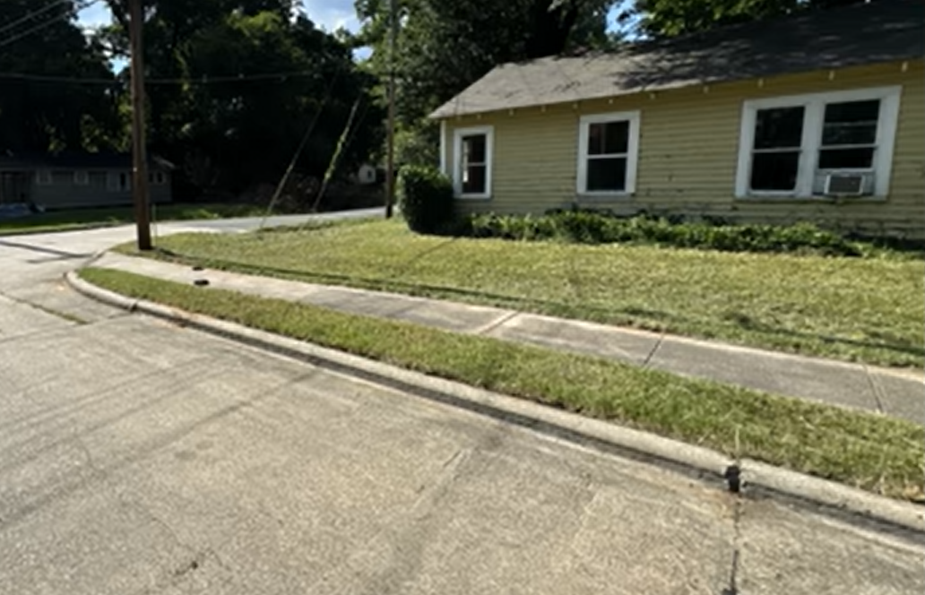 A yellow house with white window frames stands behind a sidewalk and a patch of mowed green lawn on a sunny day.