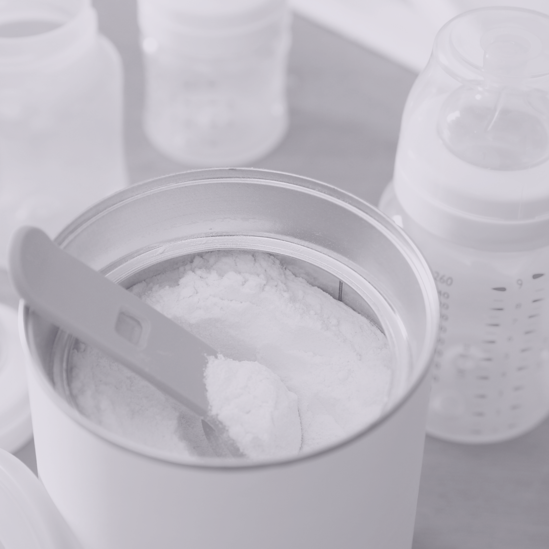 A white container of baby formula powder with a plastic scoop, next to empty baby bottles.