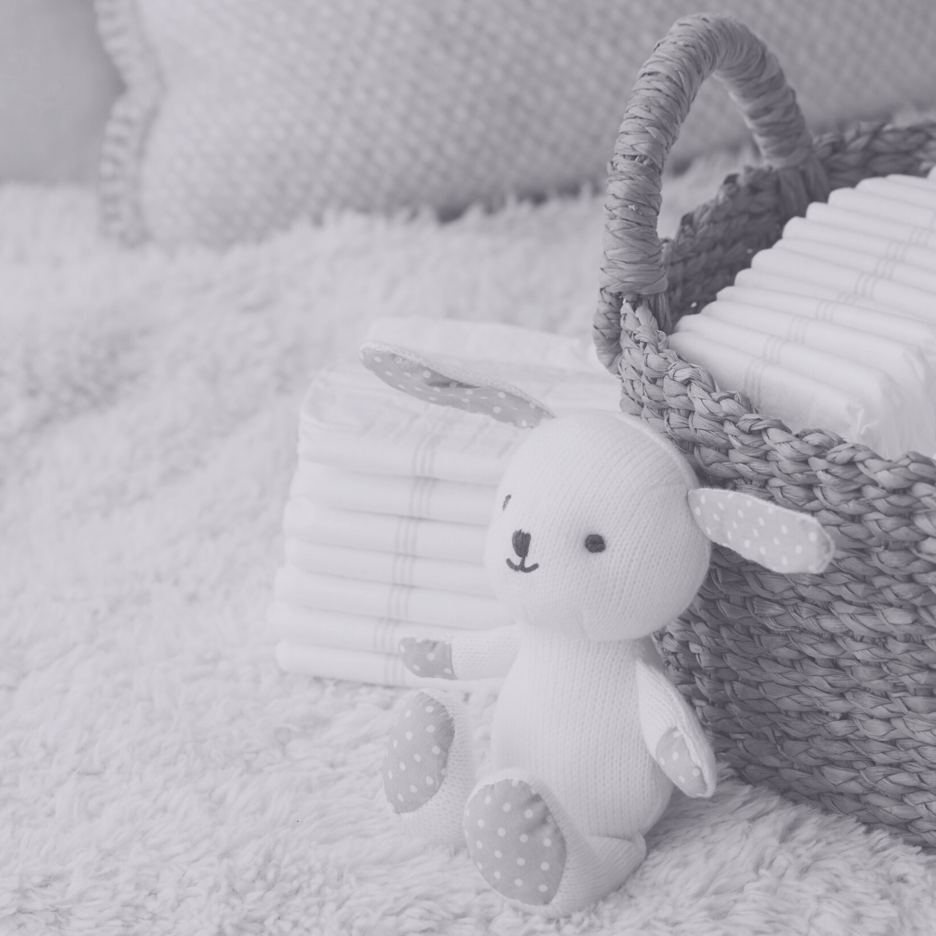 A stuffed white bunny toy sits in front of a woven basket filled with diapers on a soft, textured white surface.