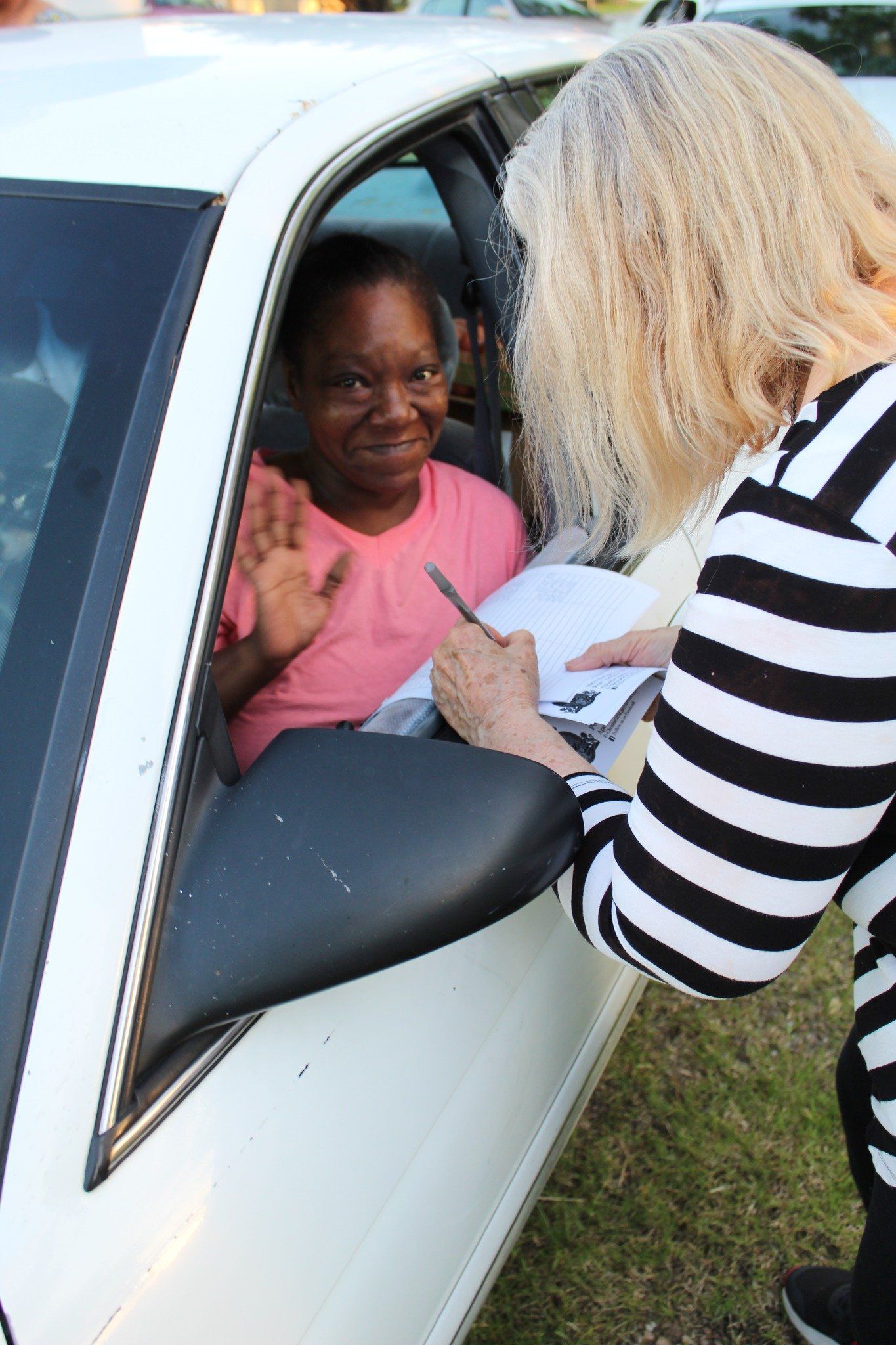 A person in a striped shirt writes on a clipboard held through the window of a white car, as the driver waves hello.