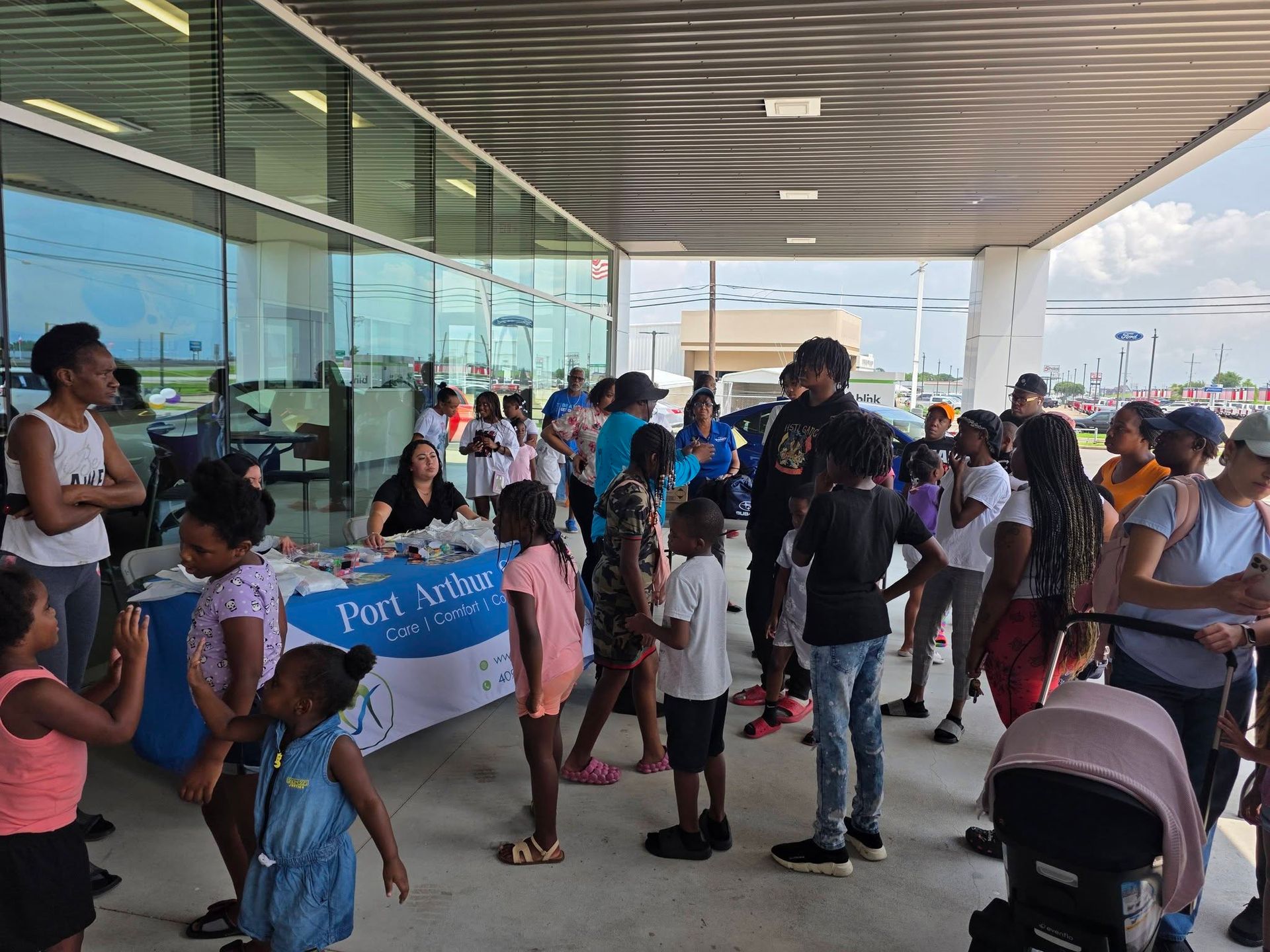 People gather at a Port Arthur community event booth under a covered outdoor walkway with glass walls.
