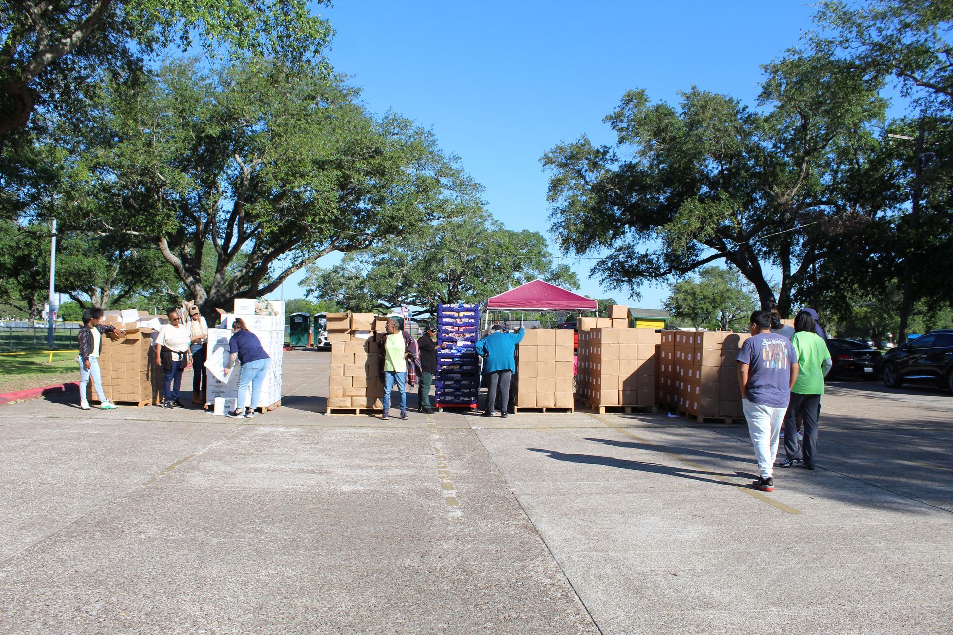 People set up rows of cardboard boxes for a community distribution event in a paved outdoor lot under sunny skies.