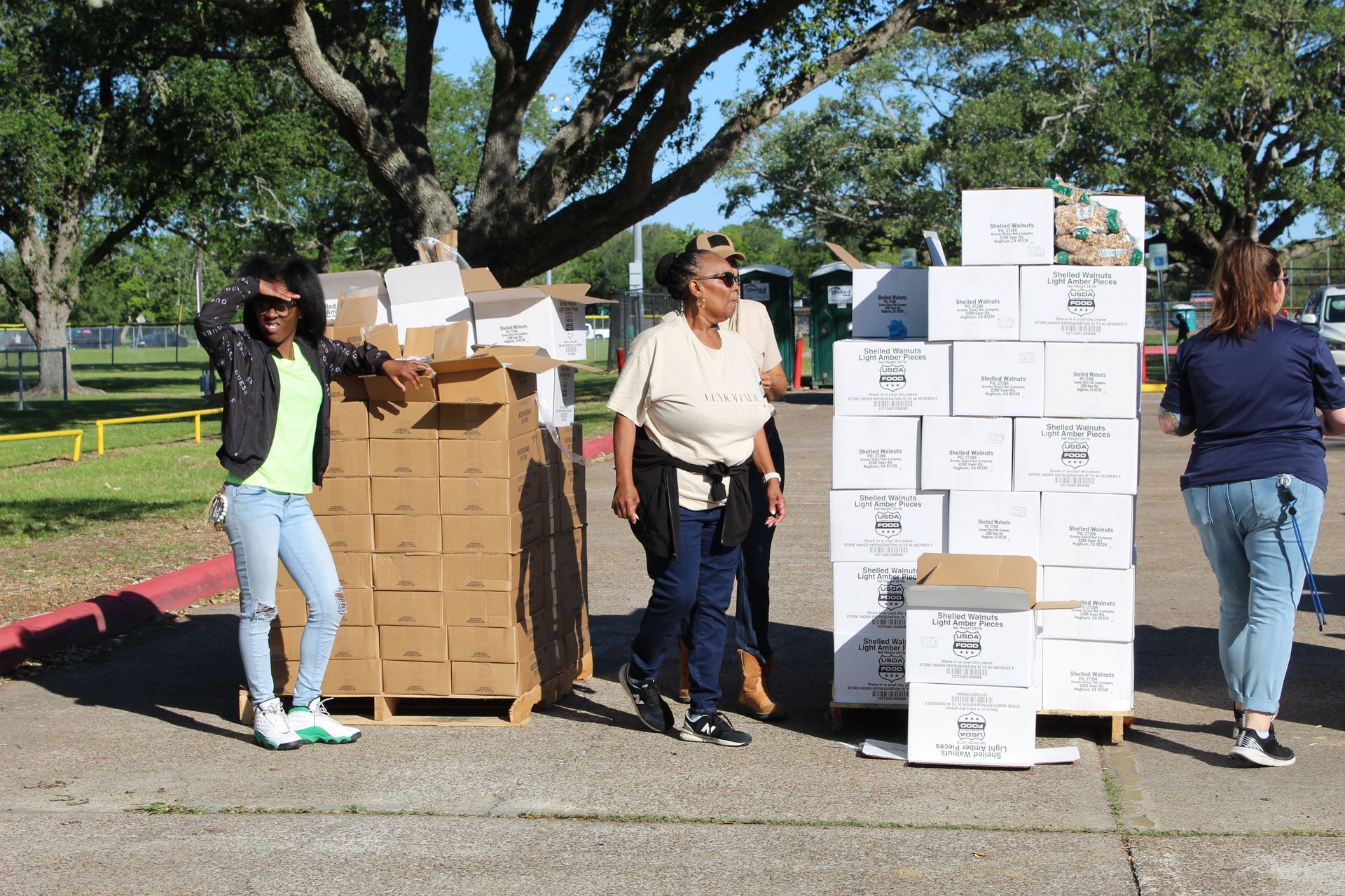 Volunteers stand outdoors beside stacks of brown boxes and white food supply crates during a community distribution event.