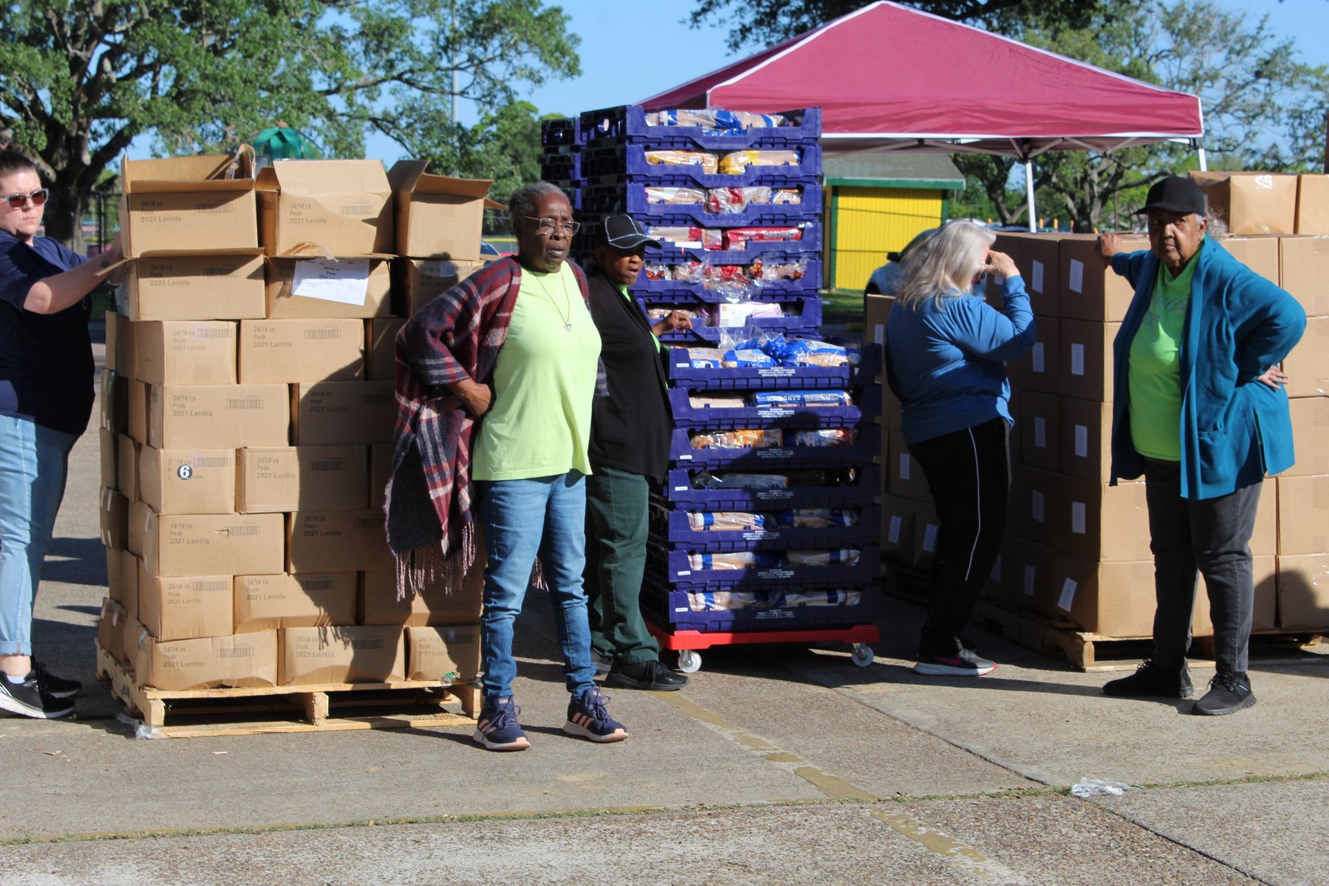 Volunteers stand outdoors by stacks of cardboard boxes and crates of food under a pink canopy.