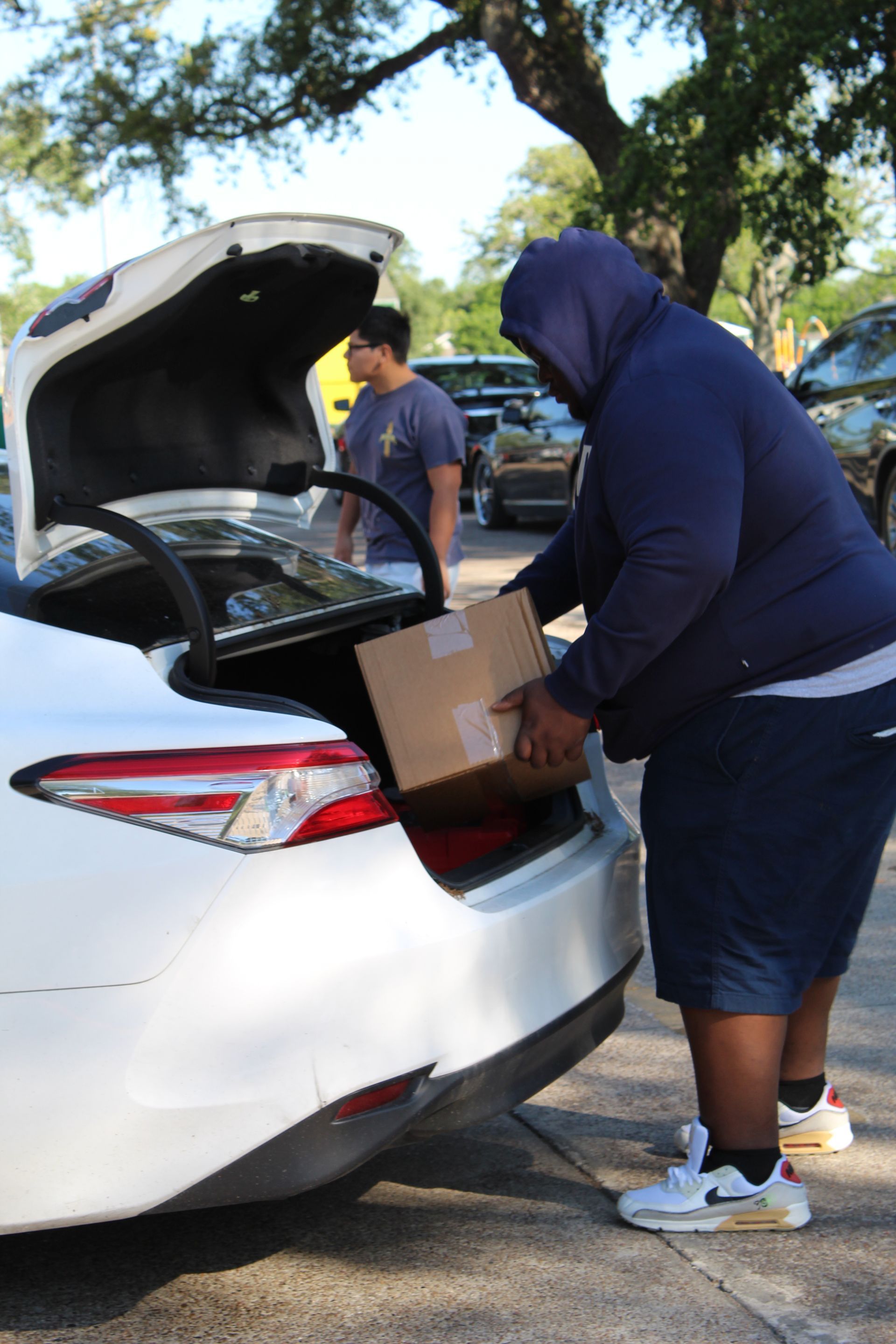 A person in a dark hooded sweatshirt places a cardboard box into the trunk of a white car in a parking lot.