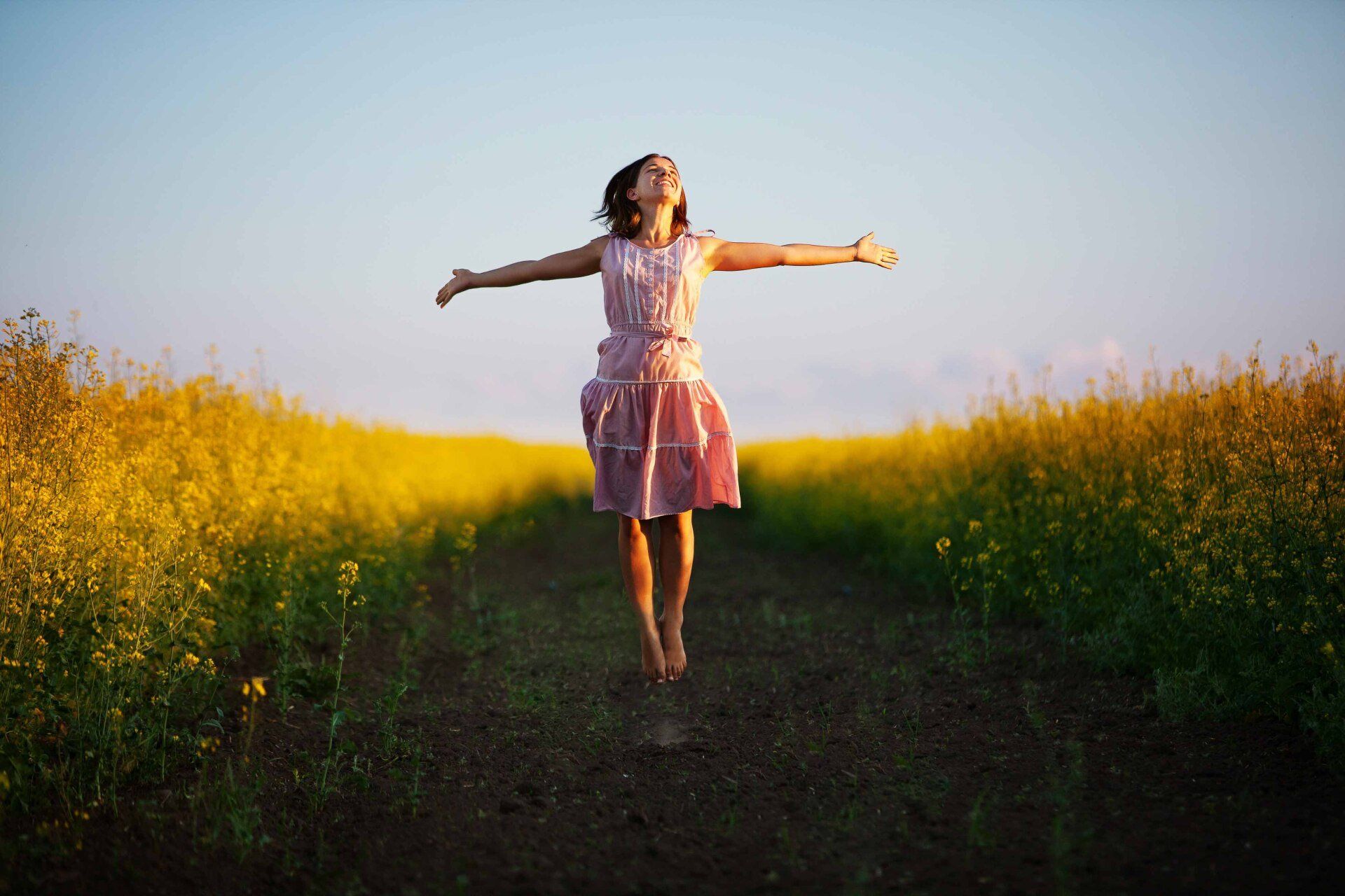Woman in pink dress with arms outstretched, leaping joyfully in a field of yellow flowers under a blue sky.